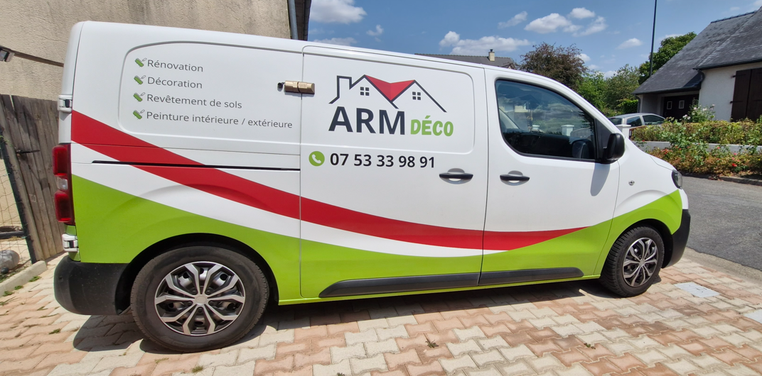 Une camionnette blanche avec une décoration verte et rouge, affichant le logo et les coordonnées d'une entreprise de décoration nommée ARMDÉCO, spécialisée en rénovation, décoration, revêtement de sols, et peinture intérieure/extérieure.