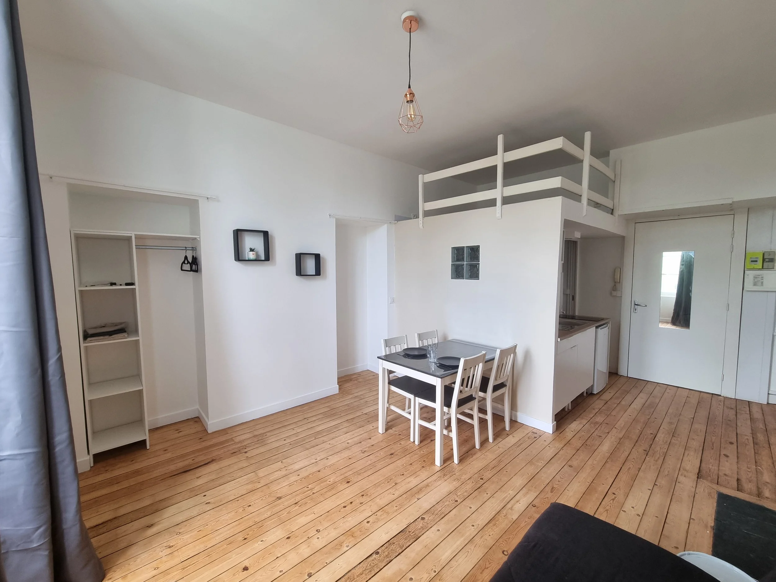 Intérieur d'un appartement avec un plafond, un luminaire suspendu, un mur blanc, une table à manger avec quatre chaises, une étagère vide, et une kitchenette avec fenêtre, au sol un parquet en bois.