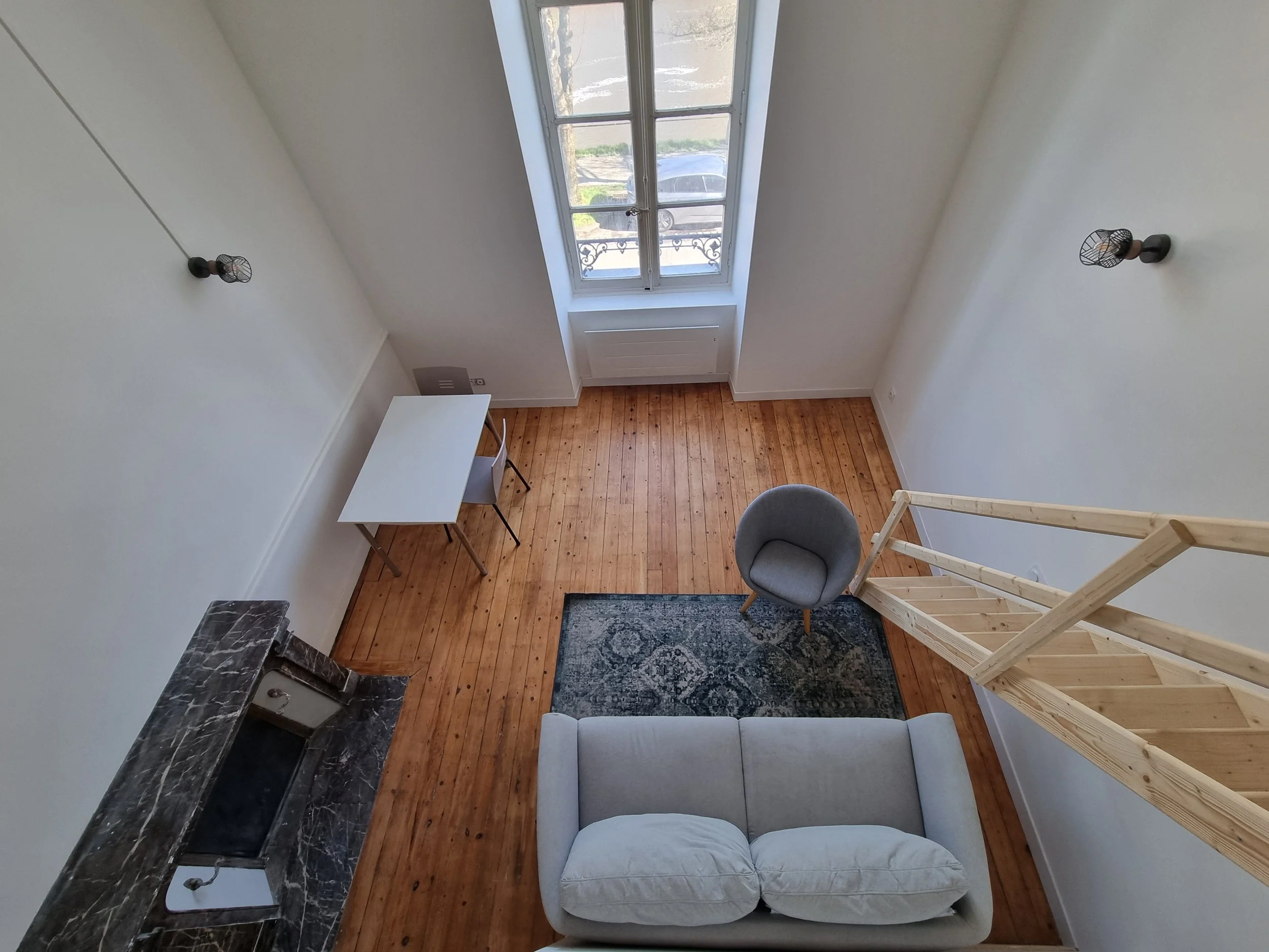 Vue d'ensemble d'un intérieur avec un plafond haut, un grand fenêtre, un fauteuil gris, un canapé gris, un tapis bleu, un comptoir en marbre noir, un bureau blanc avec deux chaises en bois, et une échelle en bois accrochée à un mur blanc.