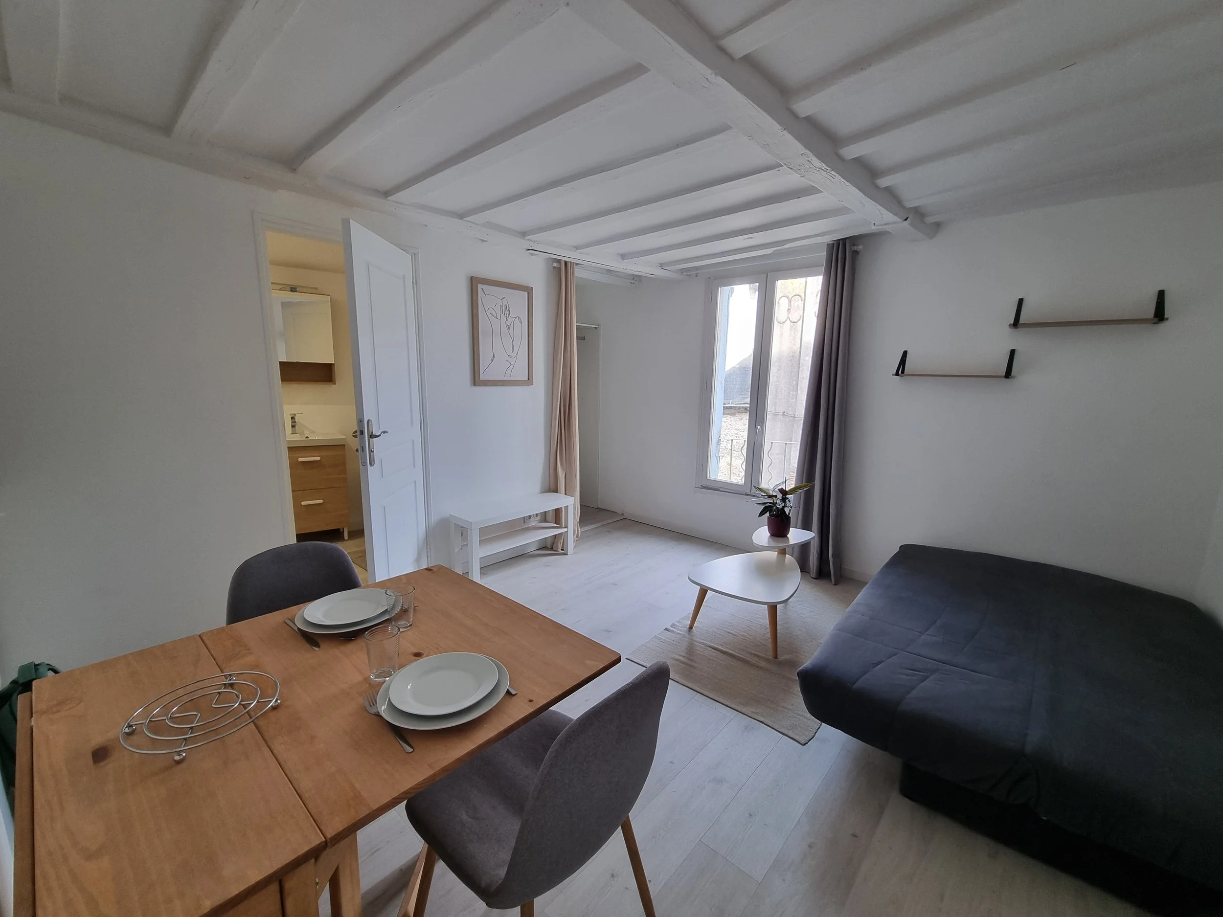 Intérieur d'un salon lumineux avec un plafond en bois peint en blanc, un canapé gris foncé, une petite table blanche, un tableau minimaliste sur le mur, et une fenêtre laissant entrer la lumière naturelle.