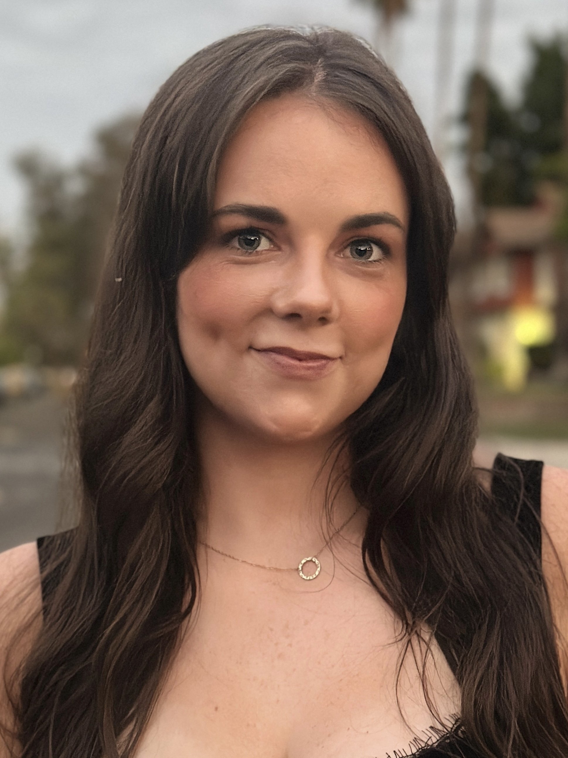 Close-up of a woman with long dark brown hair, wearing a black sleeveless top and a gold necklace with a circular pendant. She is smiling softly and the background is blurred.