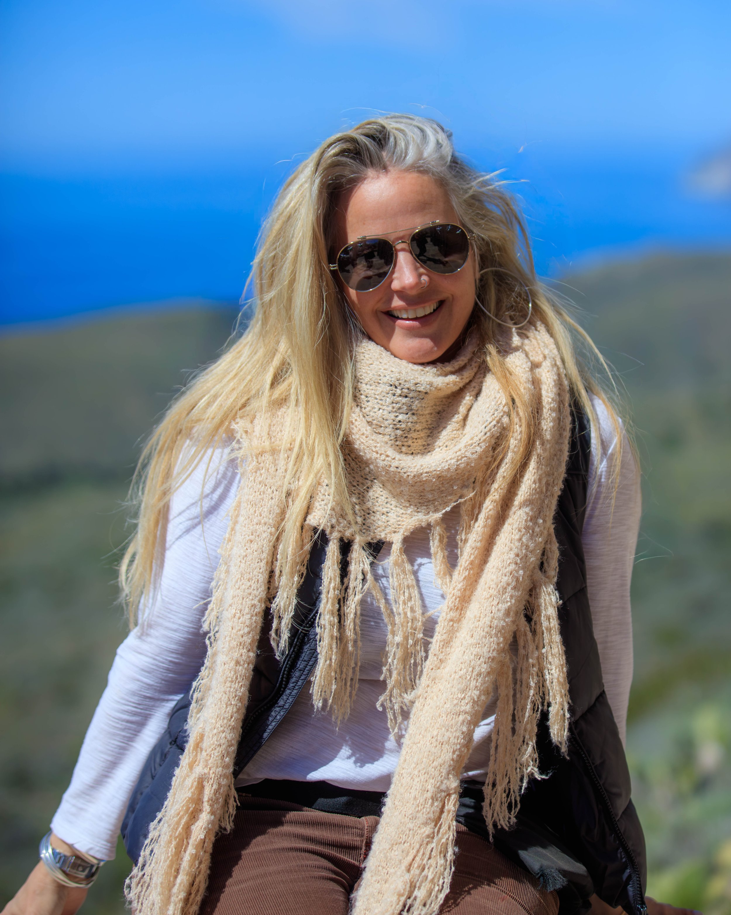 A smiling woman with long blonde hair, wearing sunglasses, a black vest, a white shirt, a light brown scarf, and brown pants, outdoors against a blue sky and green landscape.