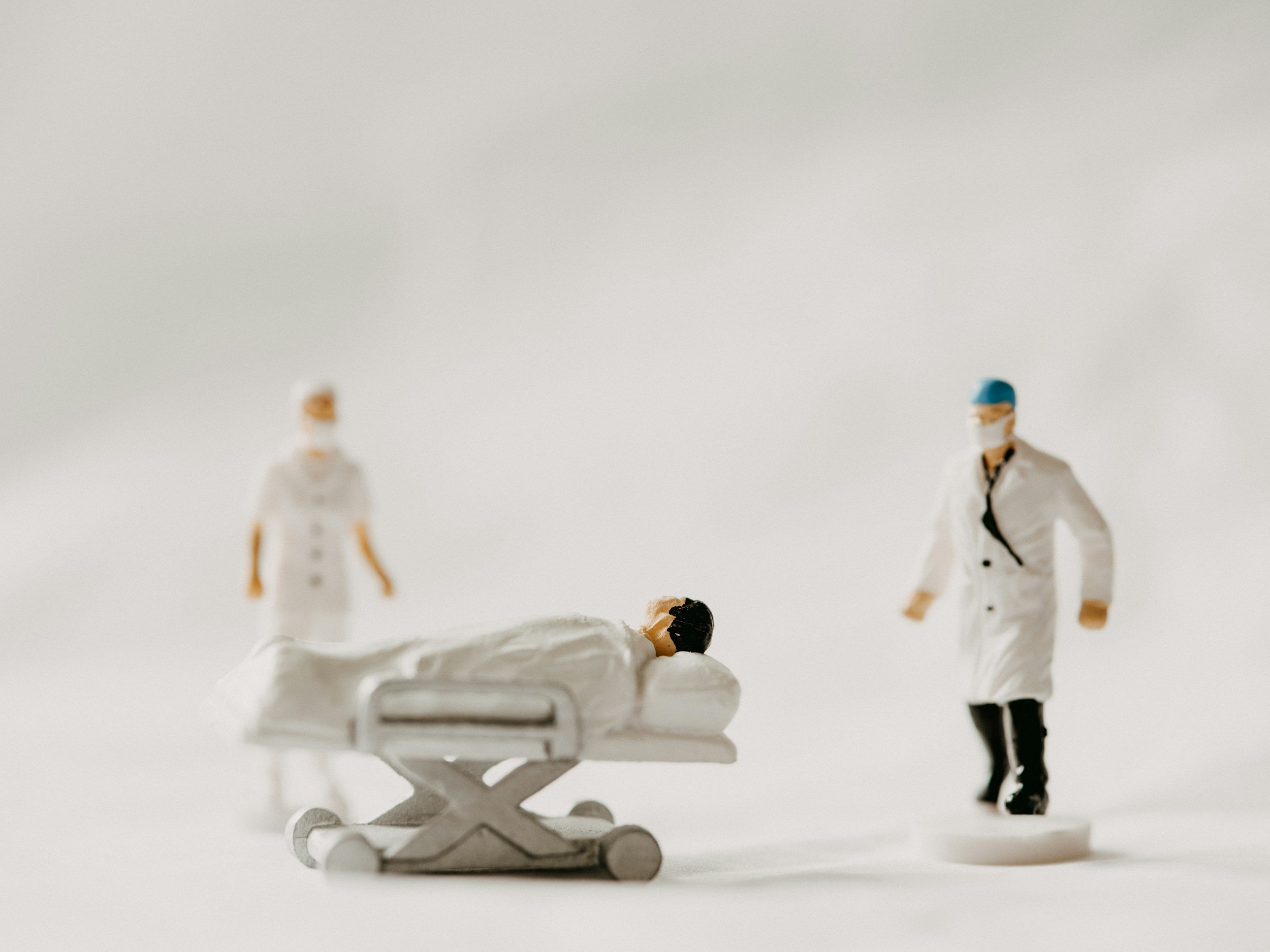 Miniaturfiguren auf einem weißen Hintergrund, eine Figur liegt auf dem Boden, zwei andere stehen, eine Person trägt eine medizinische Maske und einen Helm.