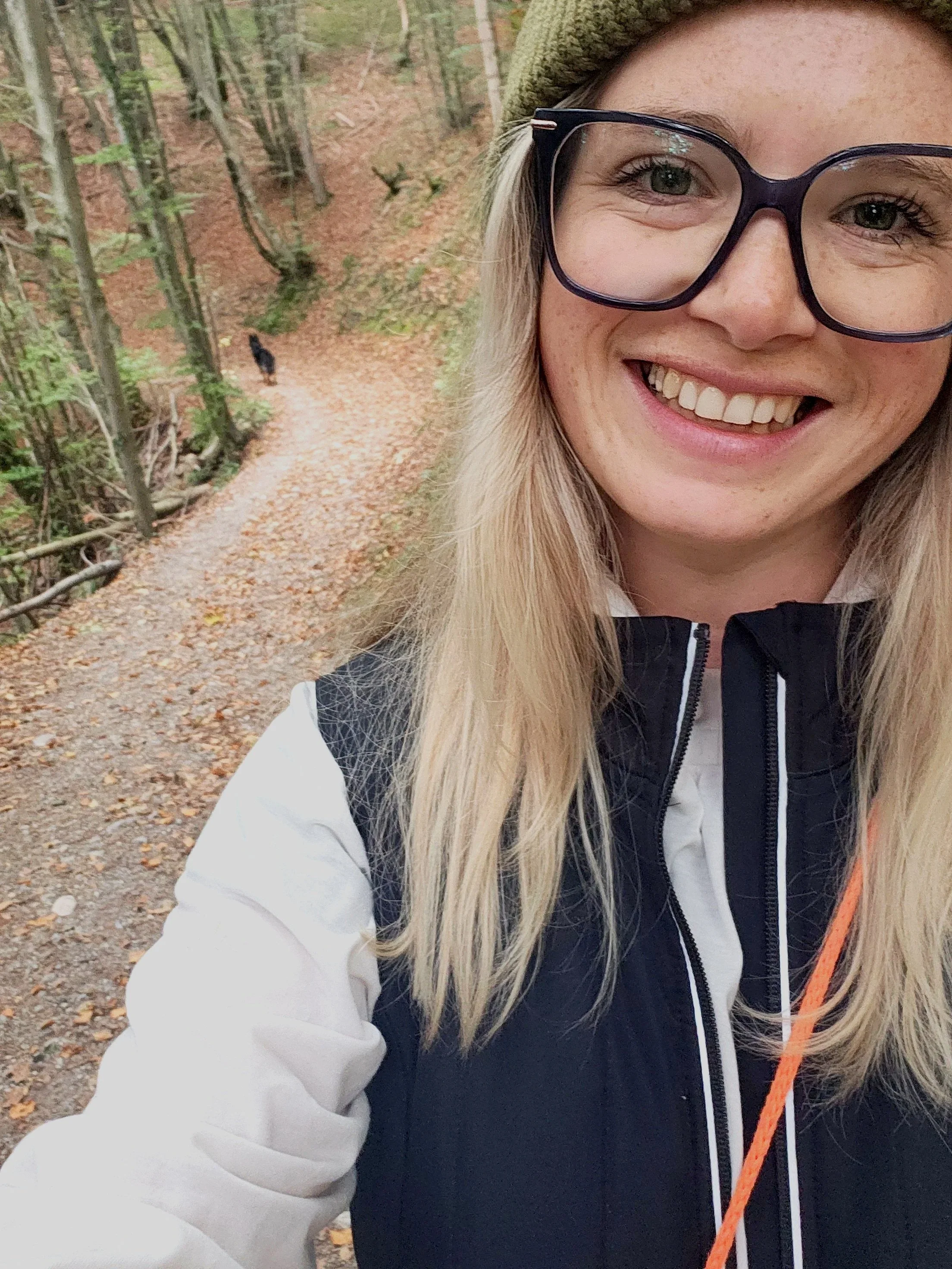 Ein lachender junger Frau mit Brille, Mütze und Jacke im Wald, in der Nähe eines Wanderweges.