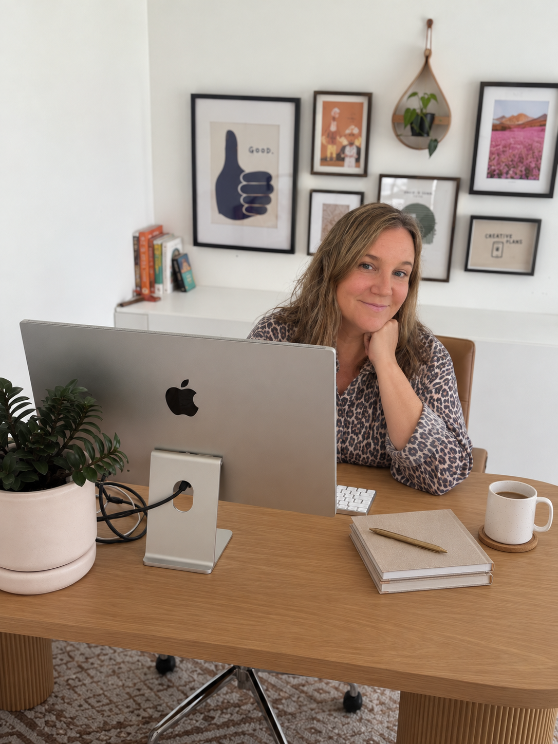 Woman working at a desk behind an Apple iMac, with notebooks, coffee, and a potted plant in a home office