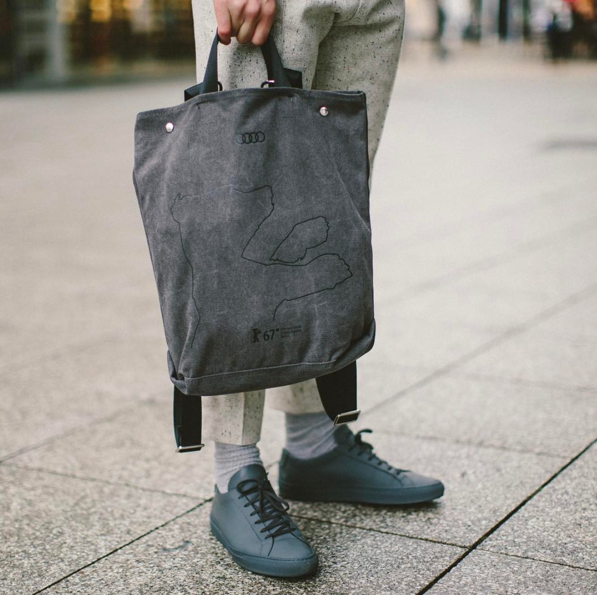 Person mit grauer Umhängetasche von Audi und grauen Sneakers auf einem gepflasterten Platz