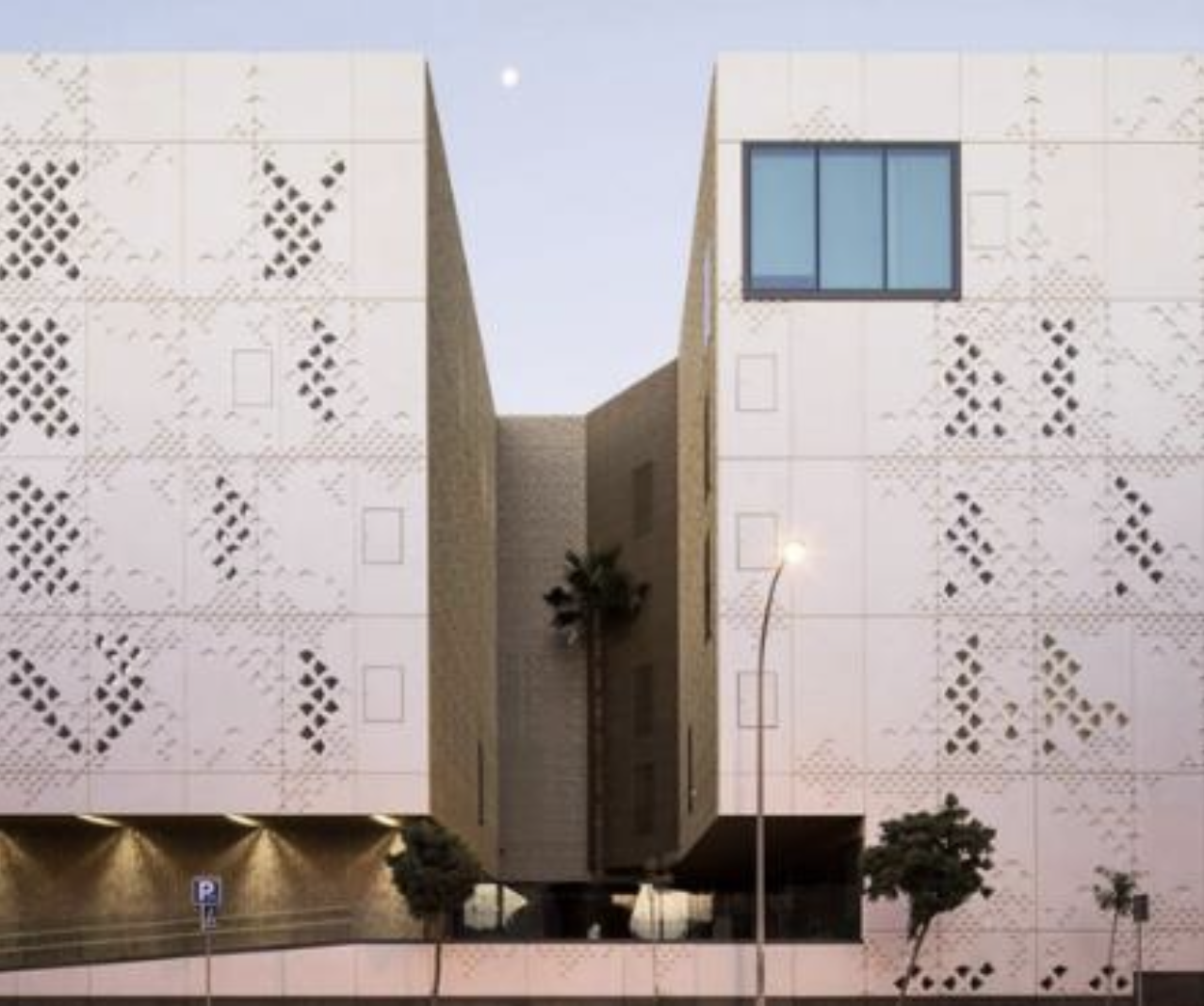 Modern building with geometric patterns on the exterior, a large window, trees, street lamp, and parking sign visible at dusk.