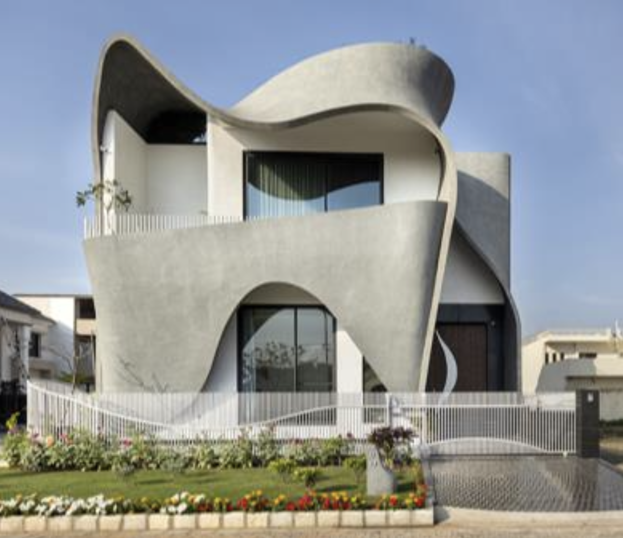 Modern multi-story house with curvy, sculptural concrete walls, large glass windows, and a white decorative fence. Well-maintained garden with colorful flowers in front.