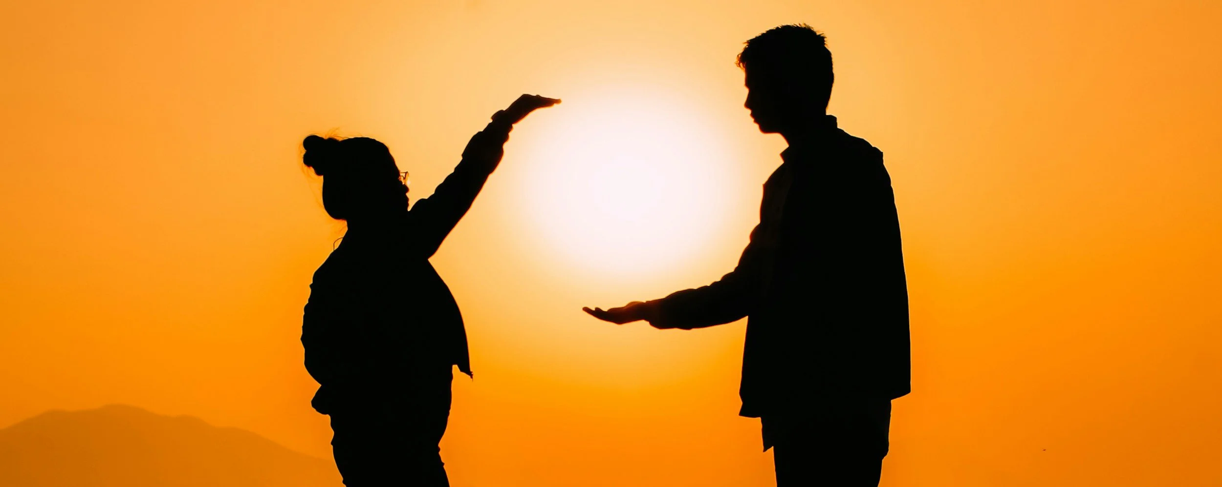 Silhouette of a girl raising her hand towards a boy, with the sun setting in the background, creating an orange sky.