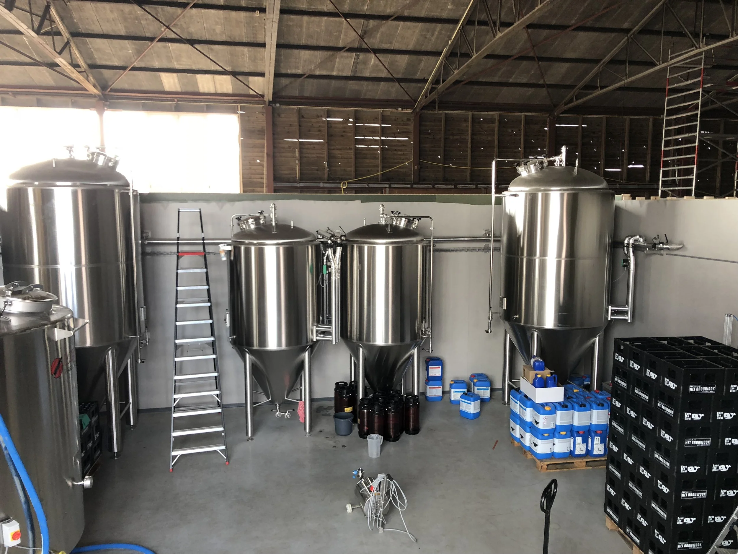 Binnen in een brouwerij met grote roestvrijstalen fermentatievaten en diverse materiaal en voorraadcontainers onder een houten plafonds met behulp van een ladder.