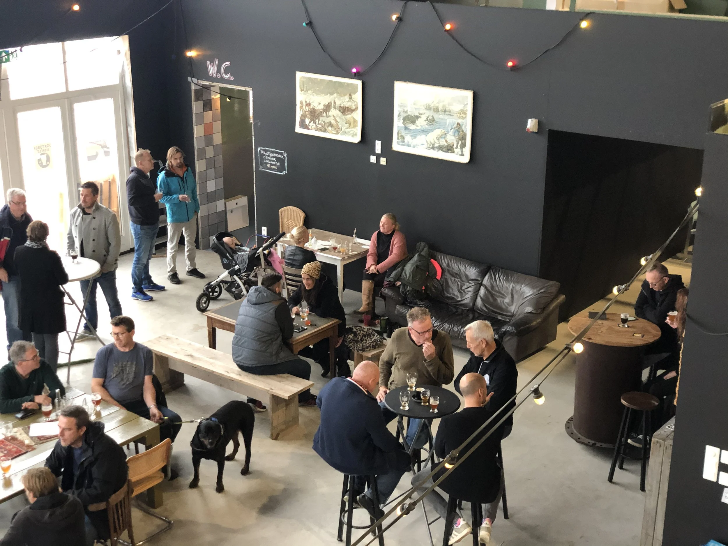 Binnenzitting van een gezellige bar of café met mensen die praten en drinken, muurschilderingen of foto's aan de muur, en kleurrijke kerstverlichting.