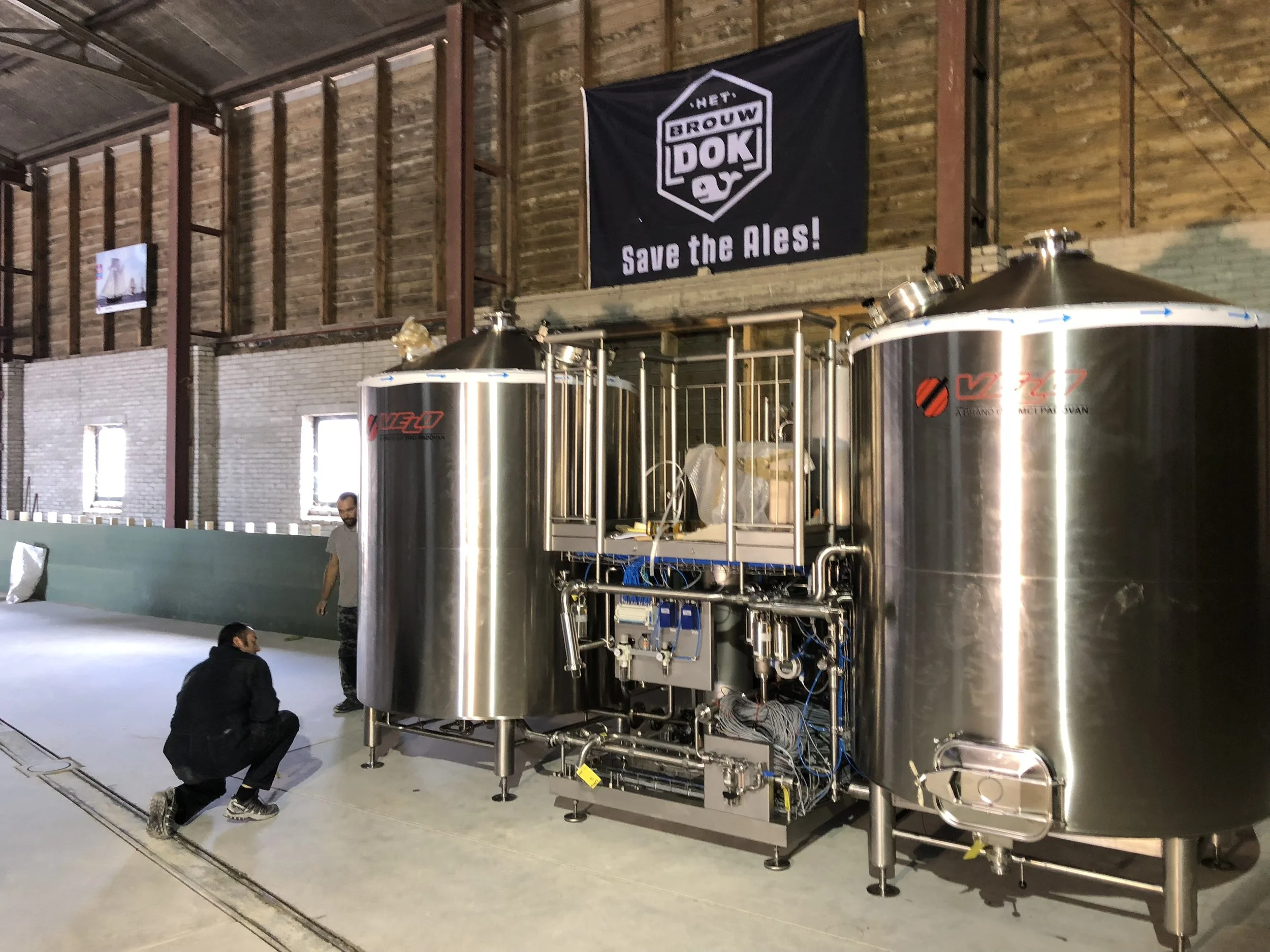 Binnen in een brouwerij, grote stalen waterketels en een banner met de tekst 'Save the Ales!', enkele mensen die de apparatuur bekijken.