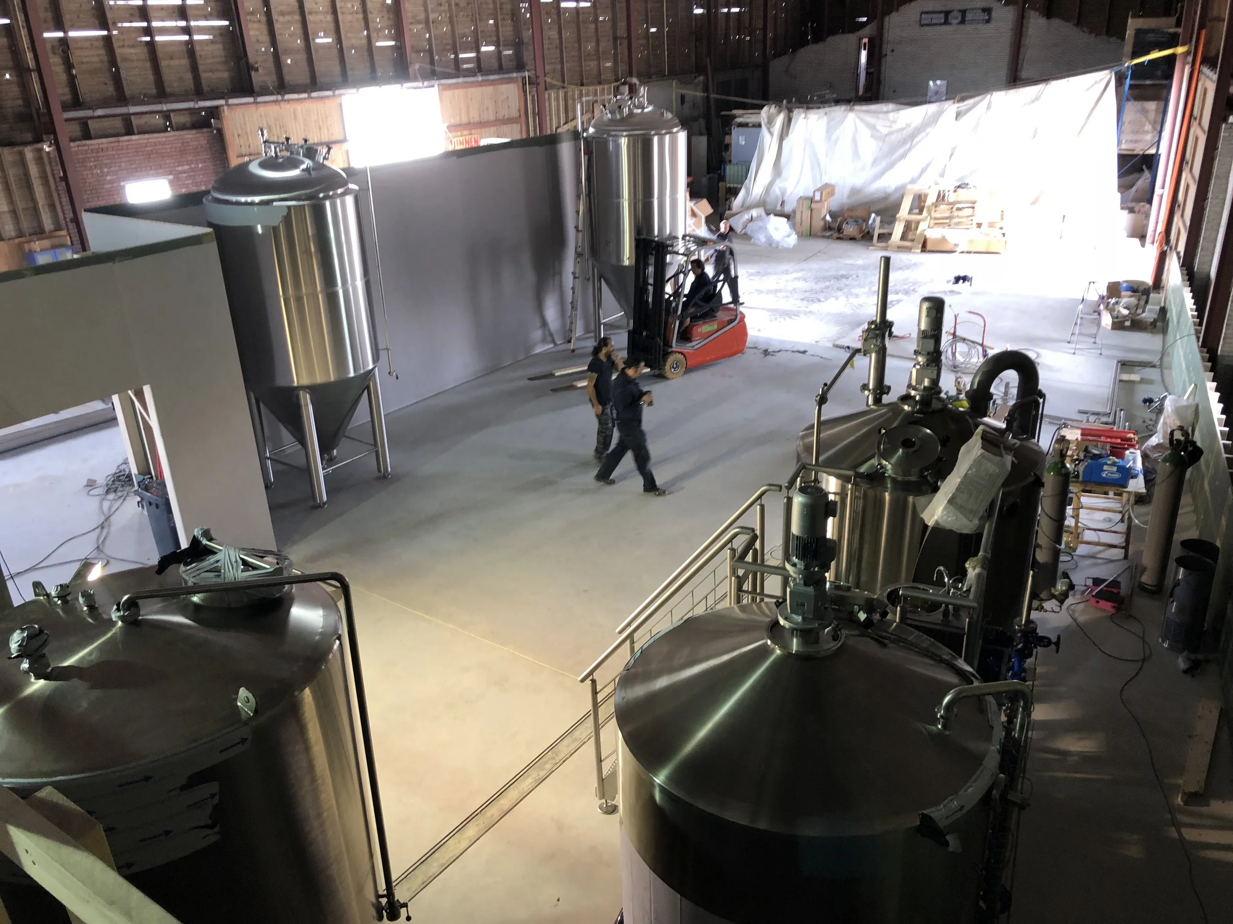 Binnenkant van een magazijn of brouwerij met grote inoxidable tanks en apparatuur. Mensen lopen in de ruimte, en er zijn verschillende machineonderdelen zichtbaar.