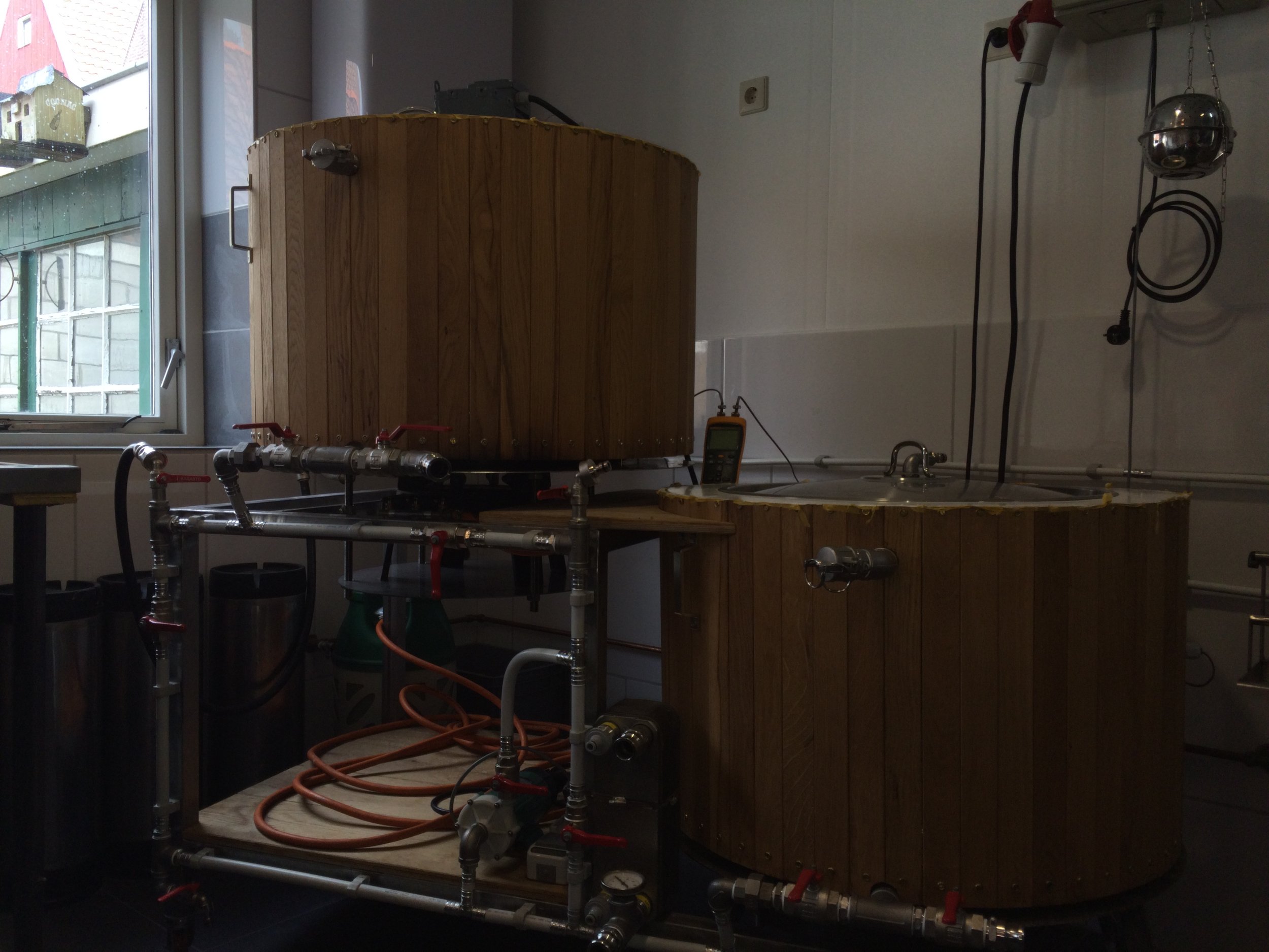 Een houten vat met een apparaat en diverse pijpen, kleppen en meters ernaast, in een keuken of werkruimte naast een raam.