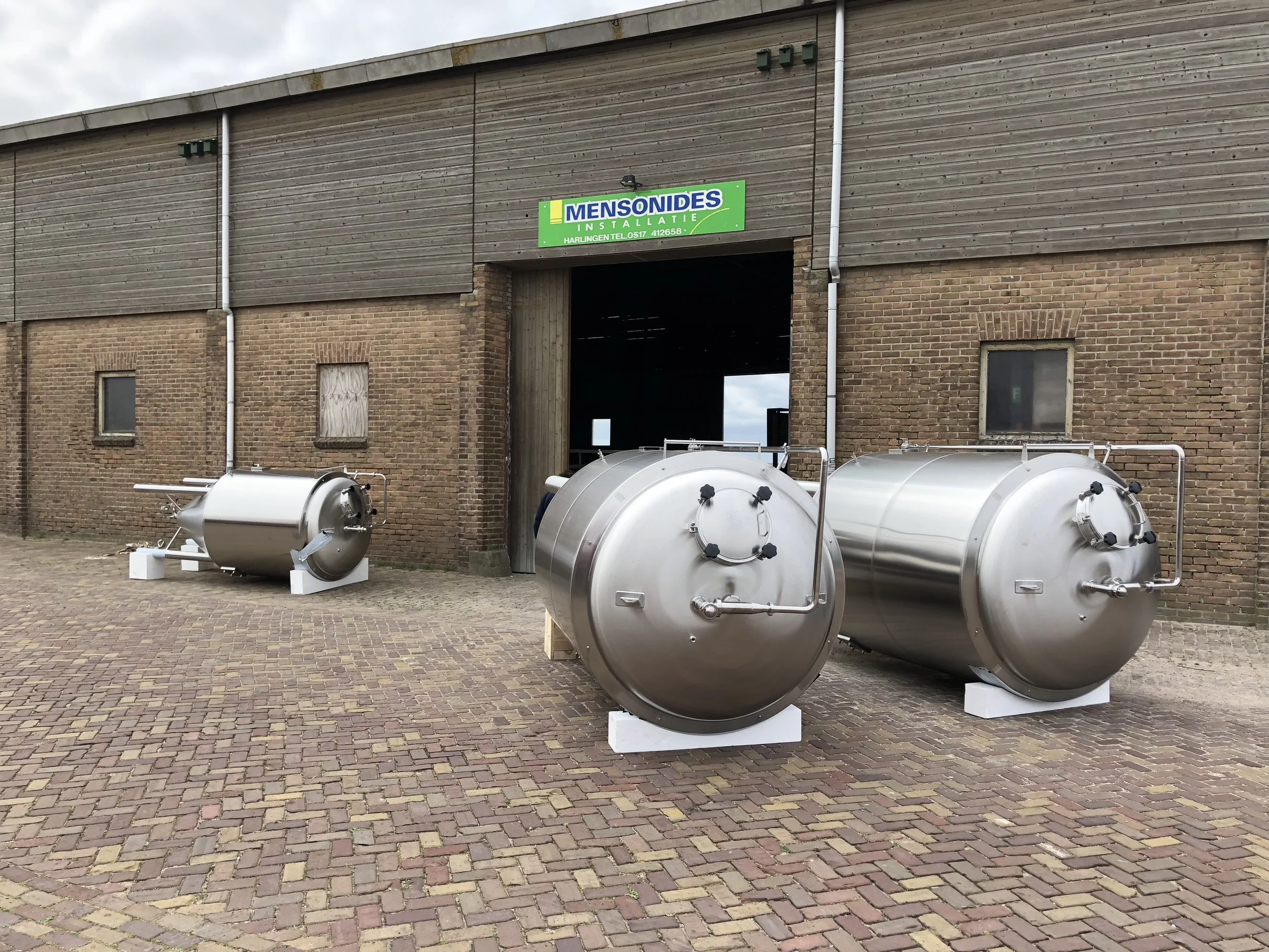Een gebouw met een houten en bakstenen gevel en een groene gevelsteen met de tekst "Mensondes Installatie". In de voorgrond liggen twee grote, metalen tanks.