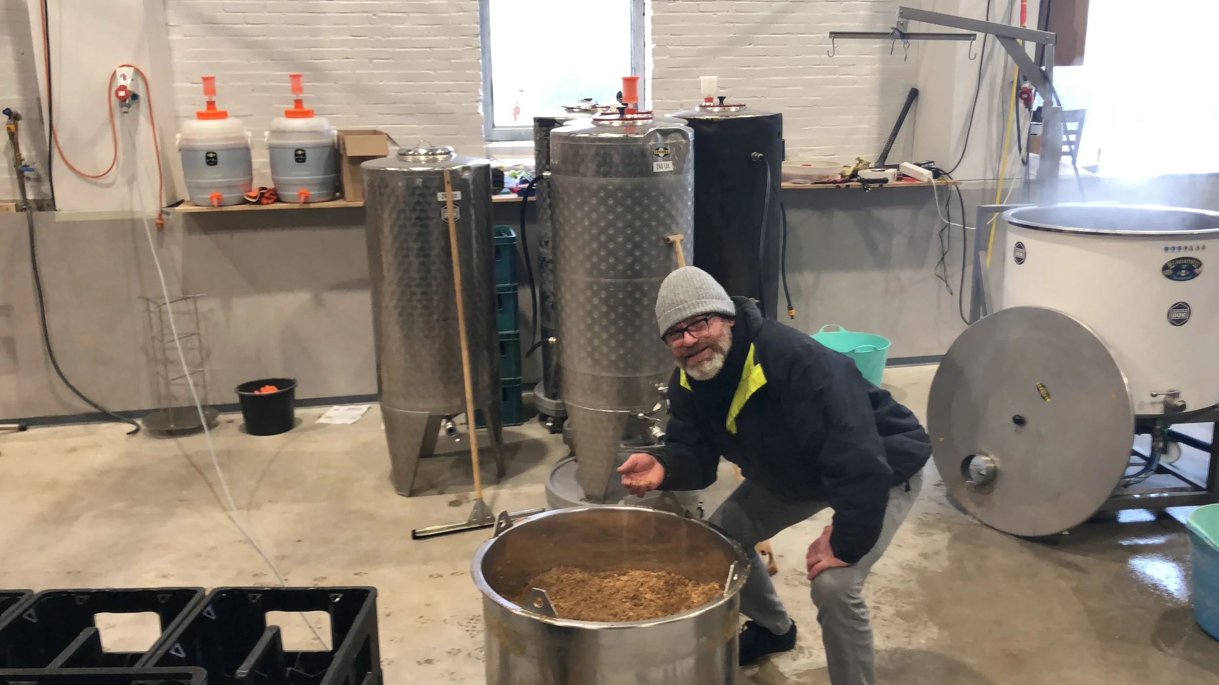 Man in een grijze muts en zwarte jas die een grote vat met gefermenteerde ingrediënten vasthoudt in een brouwerij of wijnmakerij met roestvrijstalen vaten en andere apparatuur op de achtergrond.