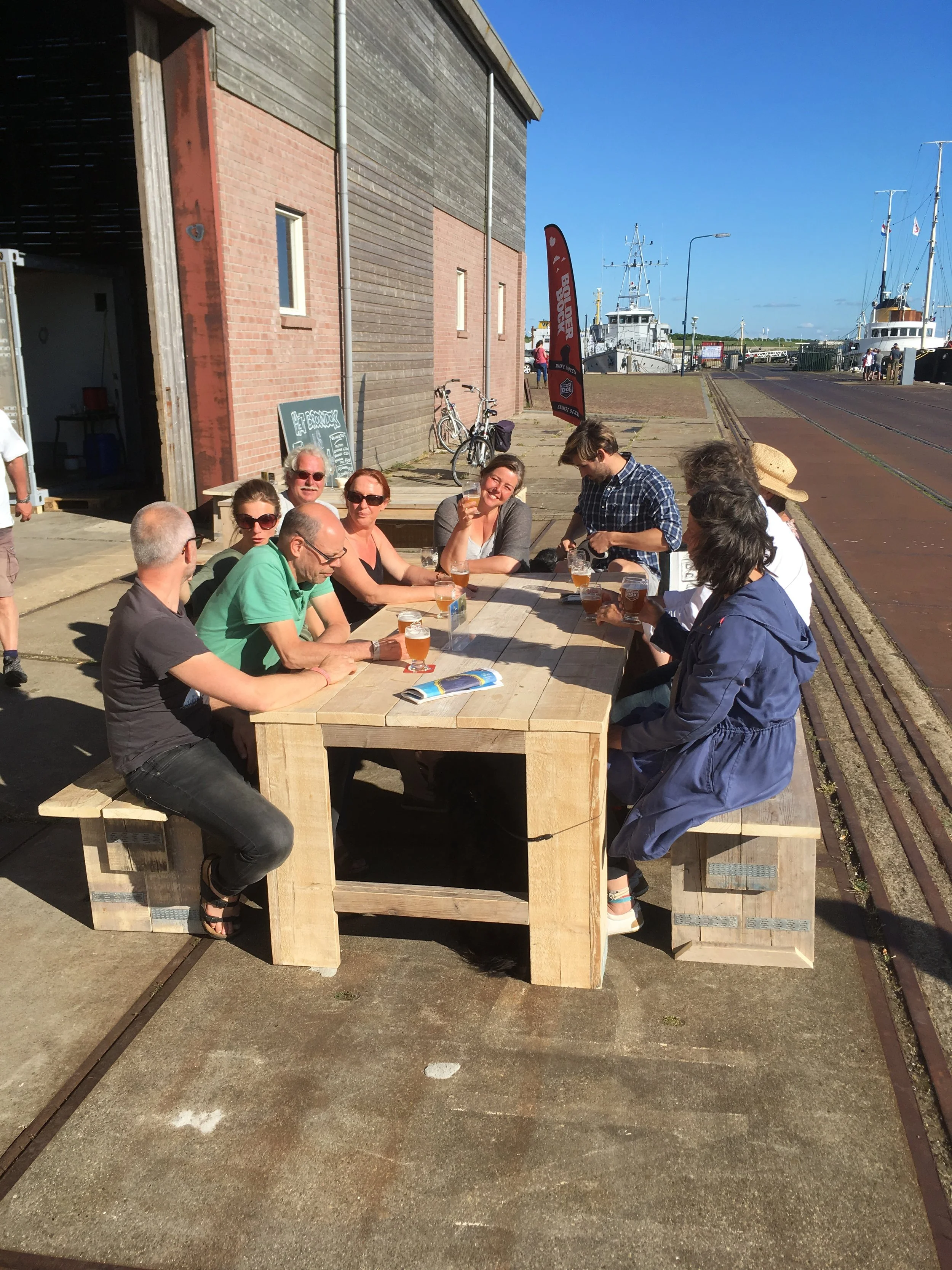 Mensen zitten aan een houten bankje en drinken bier, bij een haven met boten en een blauwe lucht.
