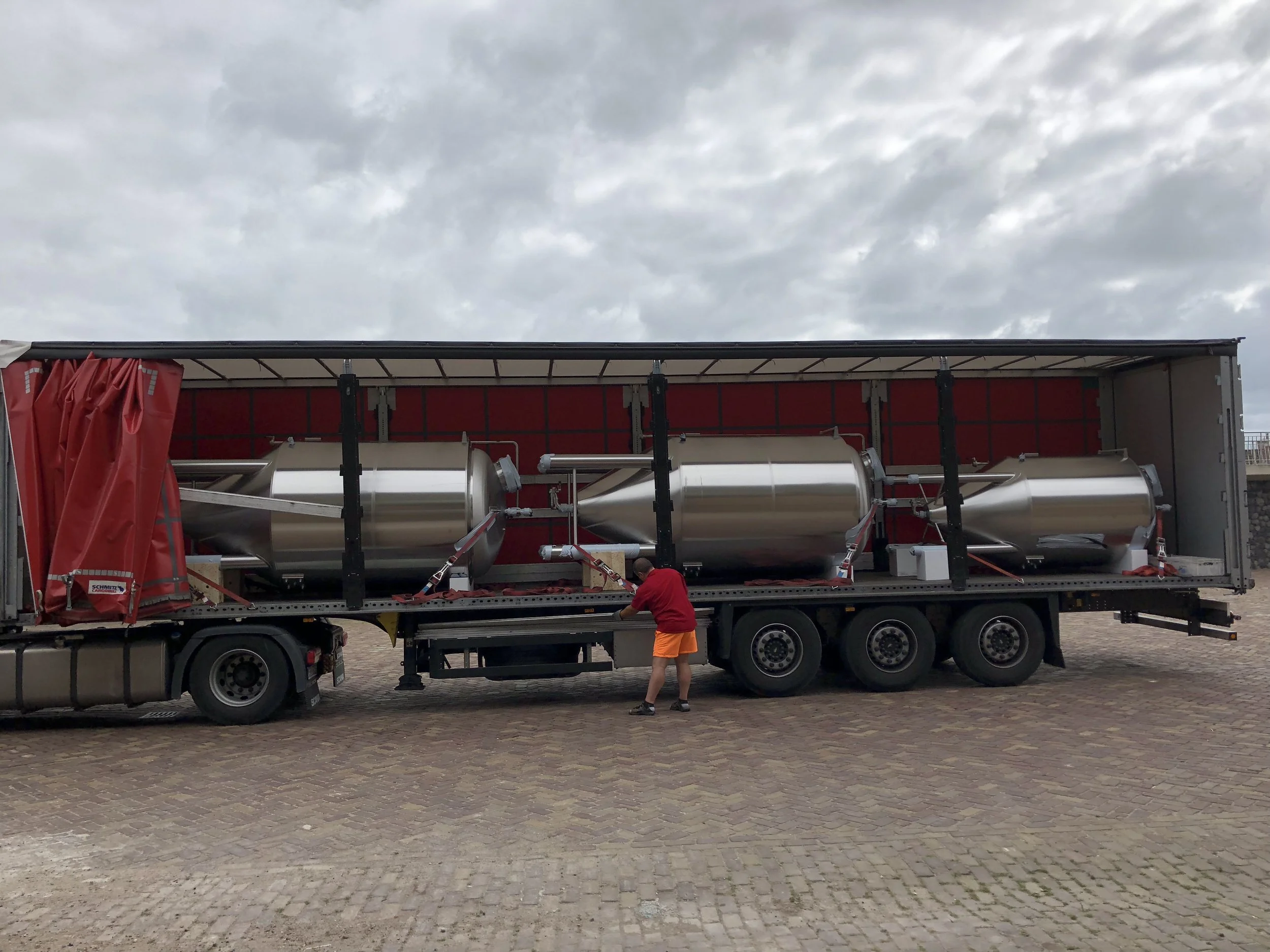 Een vrachtwagen met een grote metalen cilinder en rode onderdelen wordt op een oprit of plein geladen of aangepast, met twee mensen die er dichtbij staan onder een bewolkte hemel.
