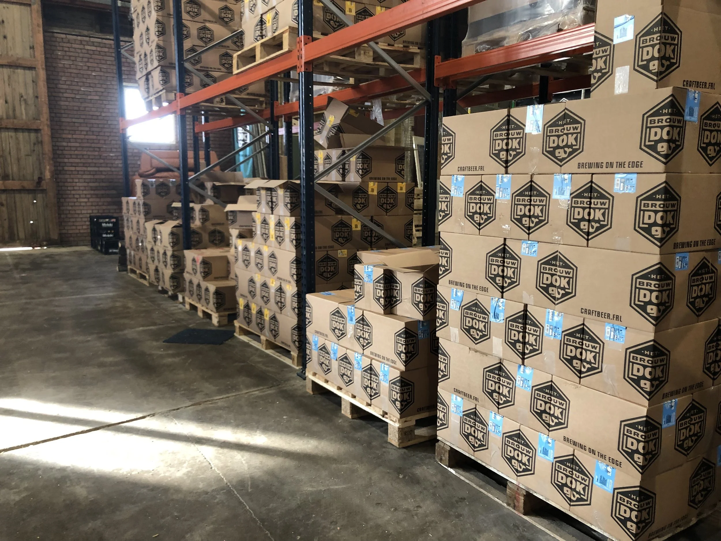 Binnen in een magazijn met houten en metalen rekken gevuld met dozen. De dozen hebben het logo van Brouwerij Het Brouw Dok en de slogan 'Brewing on the edge'.