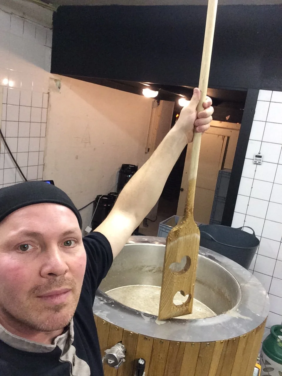 Man houdt een grote houten paddle boven een forse koperketel met een vloeistof, mogelijk in een keuken of brouwerij.