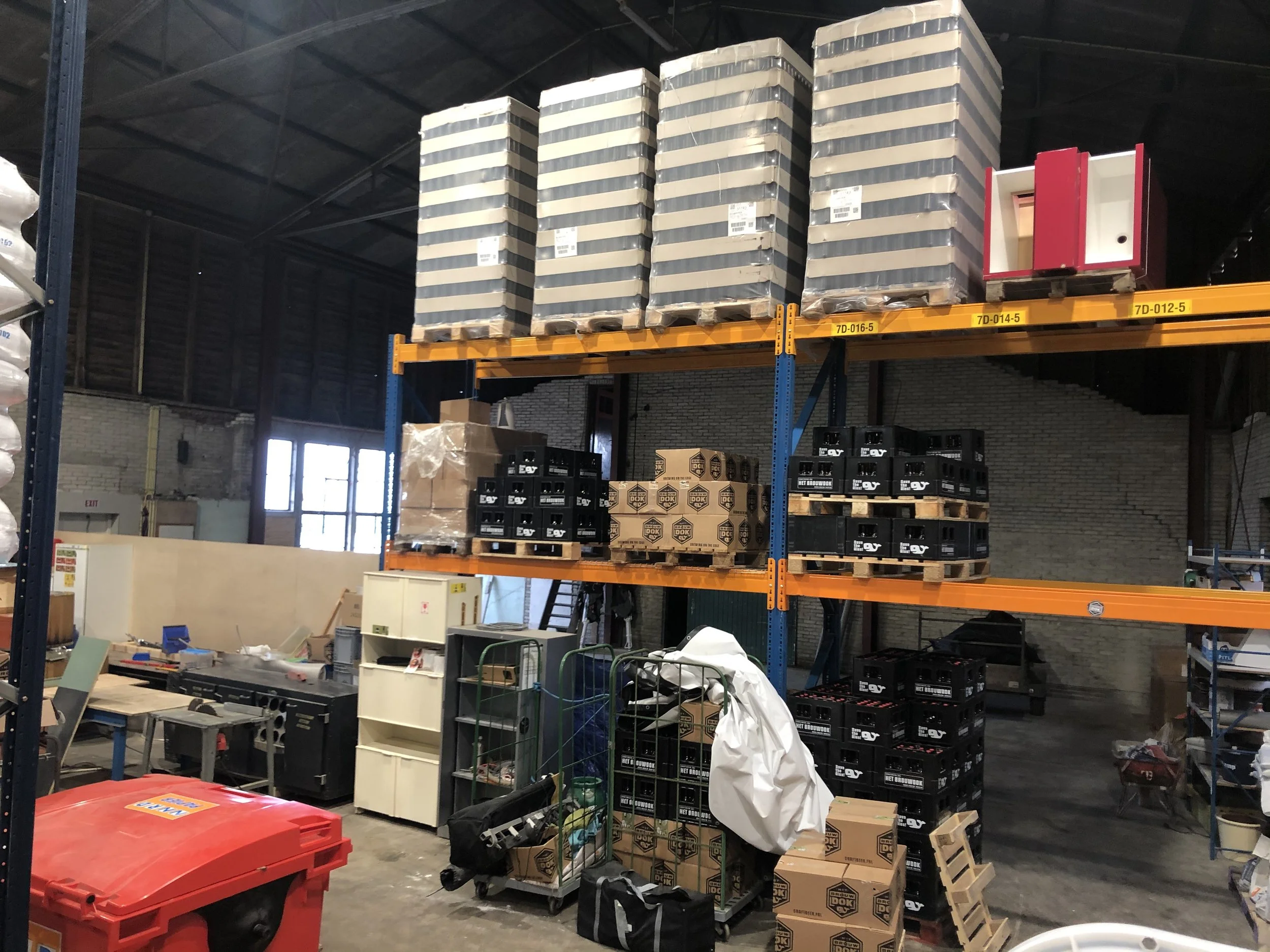Binnen in een opslagruimte met gestapelde pakken en dozen, waaronder zwarte kisten met labels en een rood kastje op een plank.