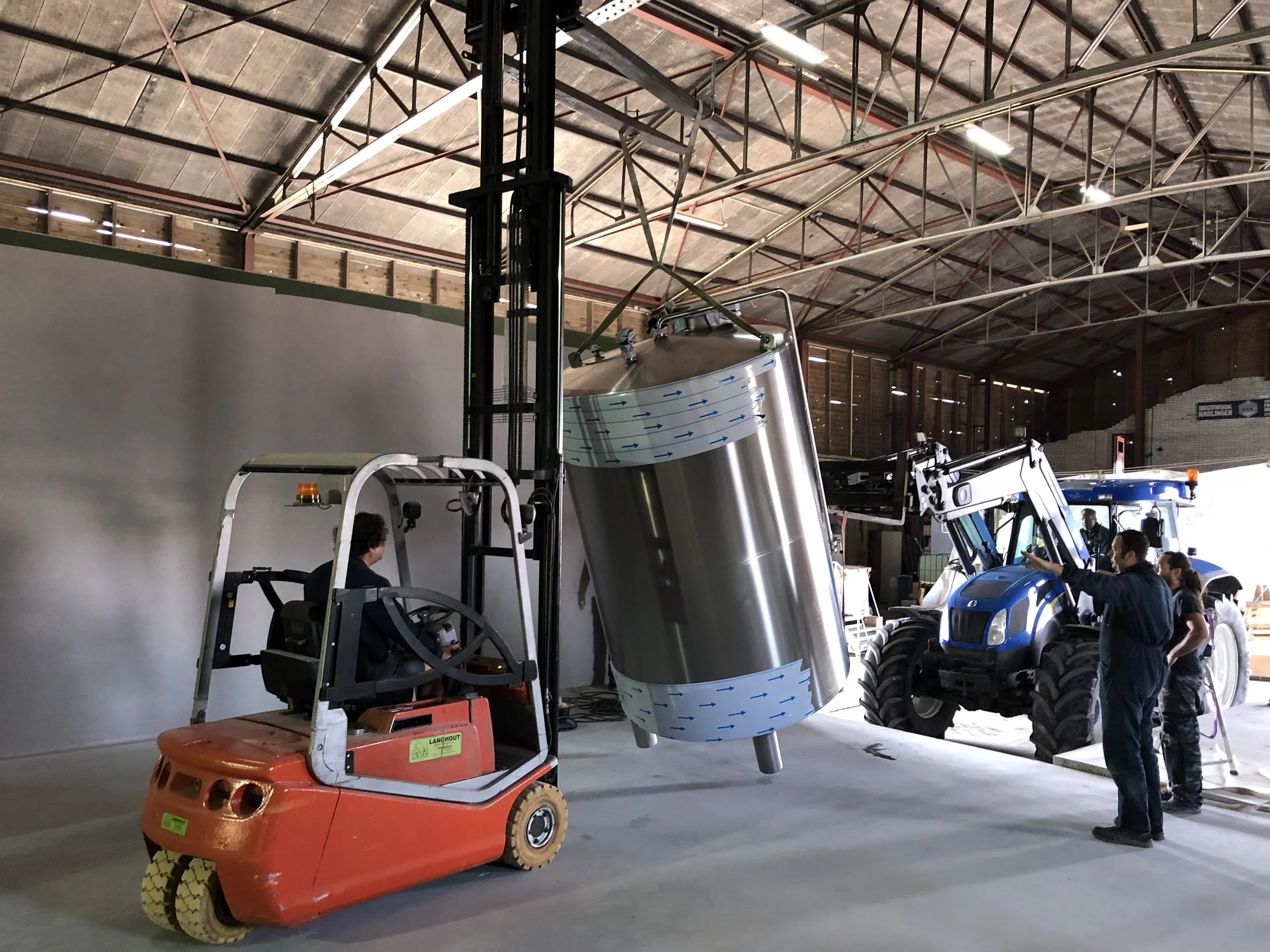 Een industriële binnenruimte met een groot metalen vat, een vorkheftruck en een tractor, omringd door een groep mensen die de machines bekijken.