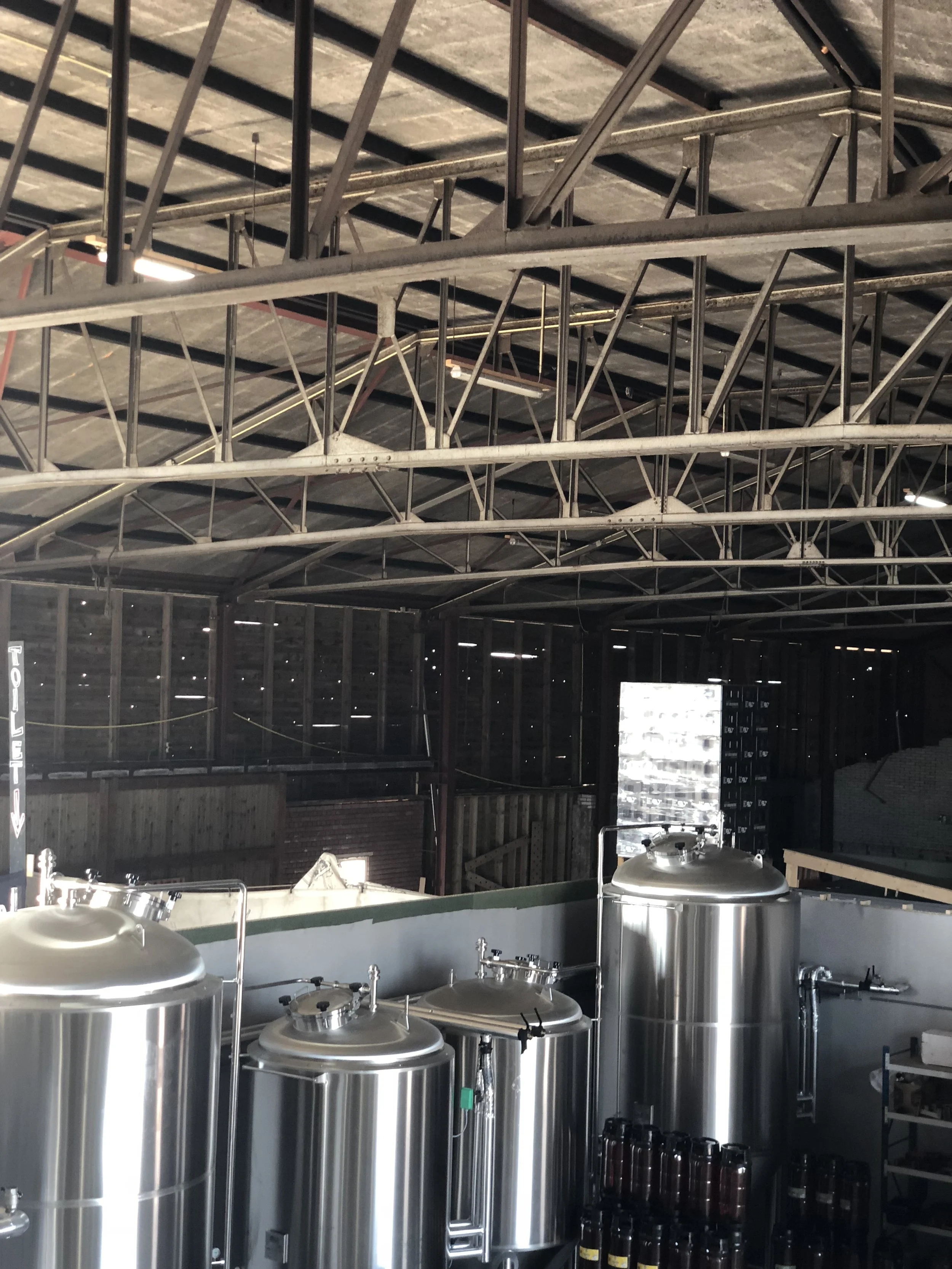 Binnenkant van een brouwerij met grote roestvrijstalen vaten en een metalen plafond met trussen.