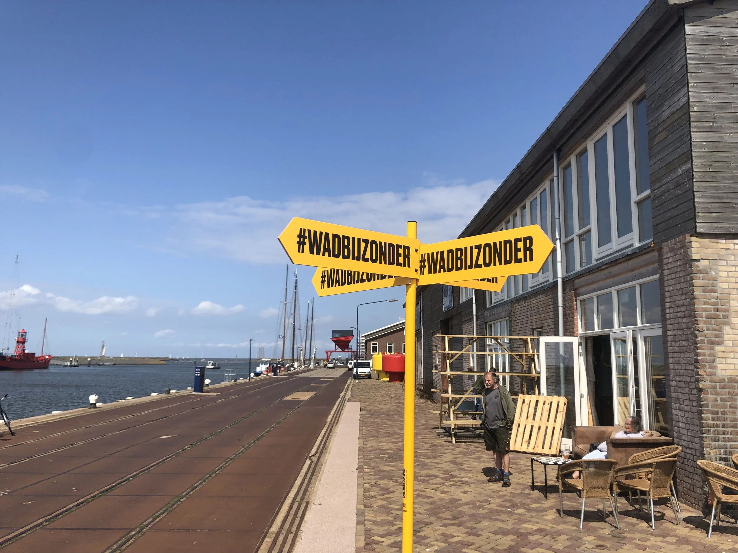 Aan de kustlijn staan gele richtingsborden met de hashtag #WADBIJZONDER, een wit gebouw met grote ramen, en mensen die daarwandelen en ontspannen.