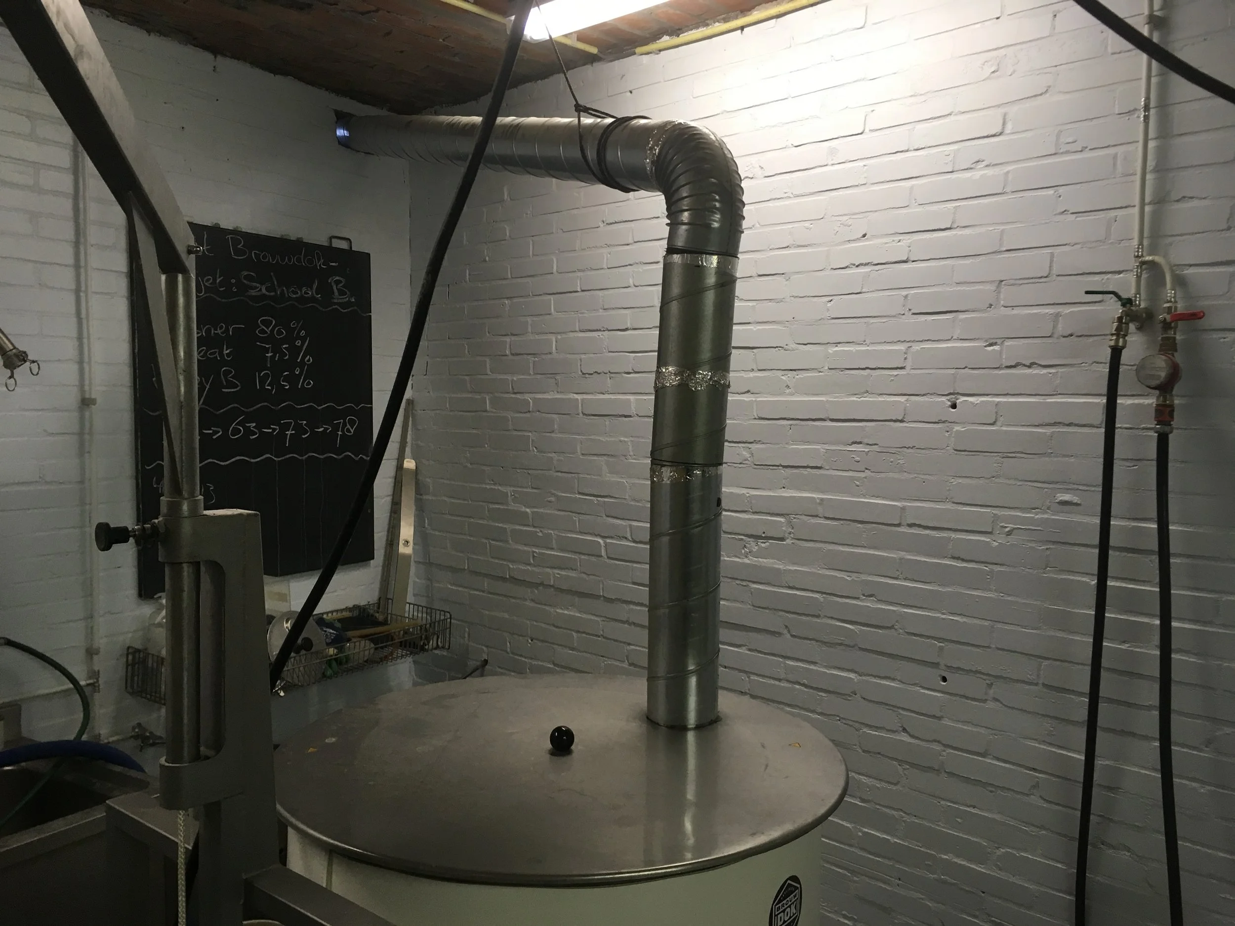 Binnenruimte met een tafel en een grote metalen buis die achter de muur uitkomt en uitsteekt. Op de muur staat een zwart bord met witte tekst. Op de vloer ligt een kleine zwarte bal.