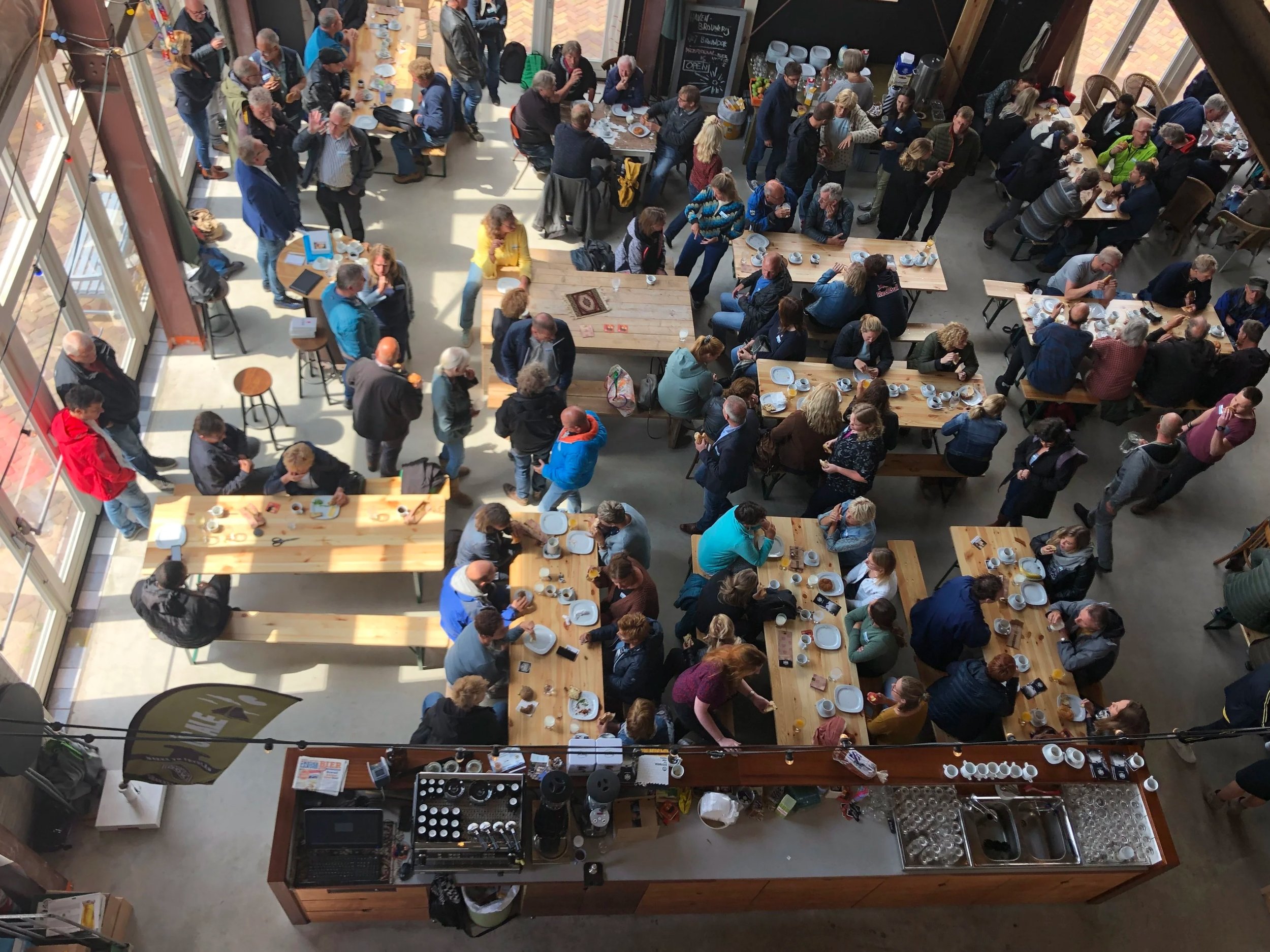 Overzicht van een druk café of restaurant vol met mensen die genieten van hun maaltijd en drinken, met houten tafels en grote ramen die natuurlijk licht binnenlaten.