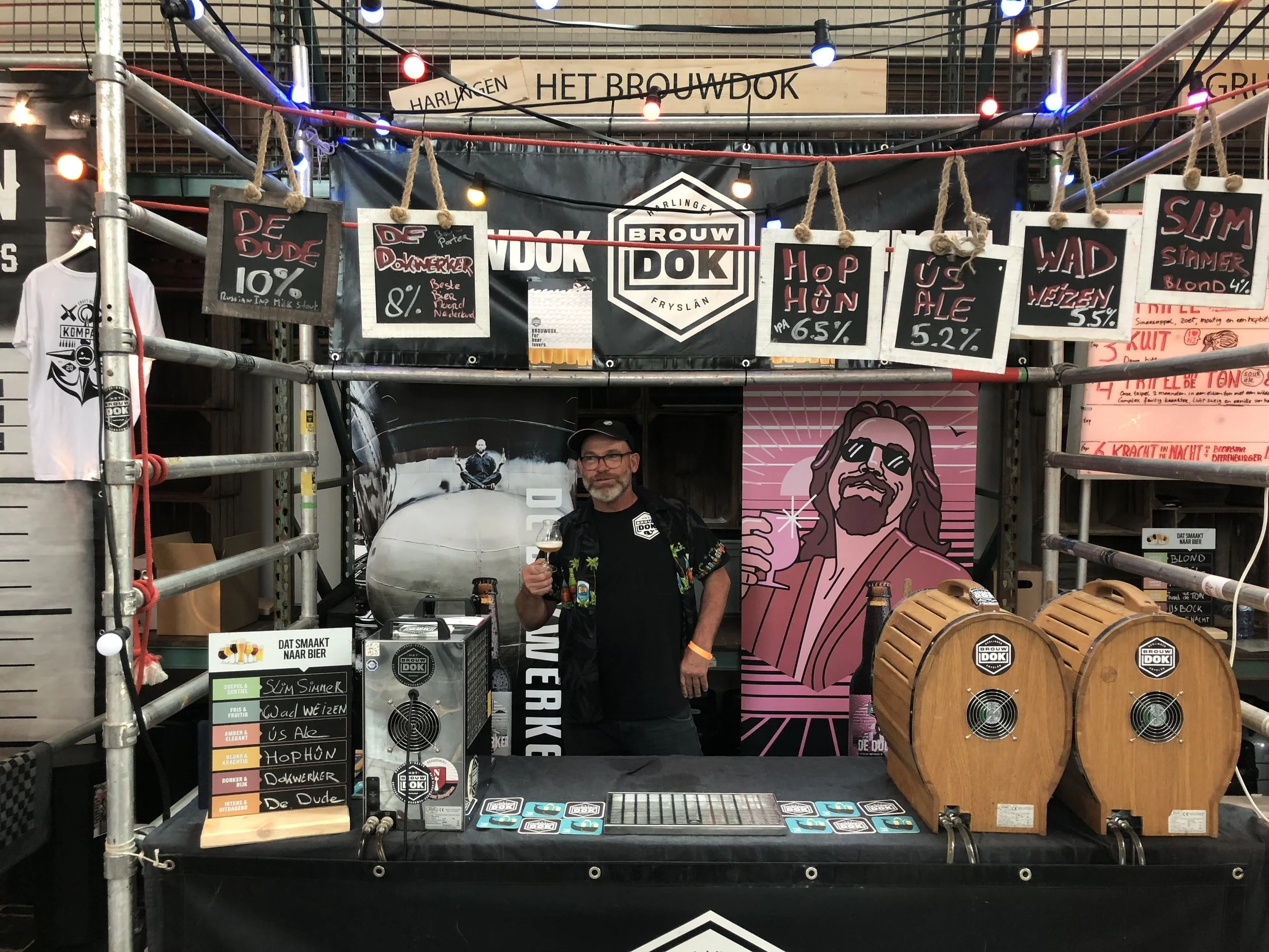 Een stand met verschillende bieren en promotiekaartjes, inclusief een man die proost met een glas bier. Er hangen meerdere borden en decoraties, waaronder een afbeelding van een persoon met dreadlocks en een overzicht van bierdetails.