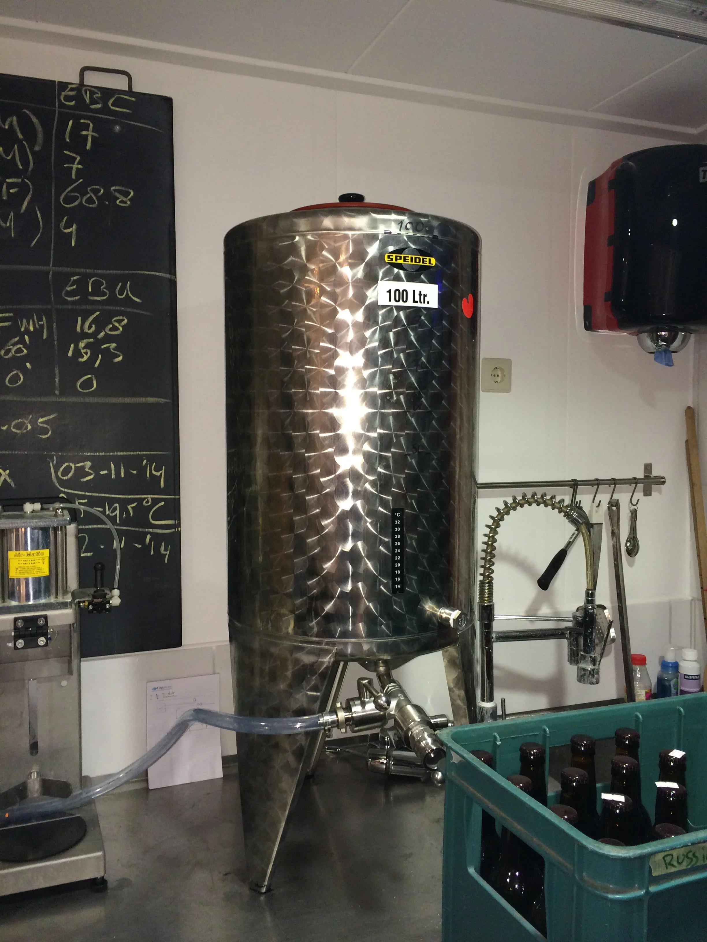 Rvs tank met een label '100 Ltr.' in een brouwerij of laboratorium, omgeven door verschillende apparatuur en een groene container met glazen flessen.