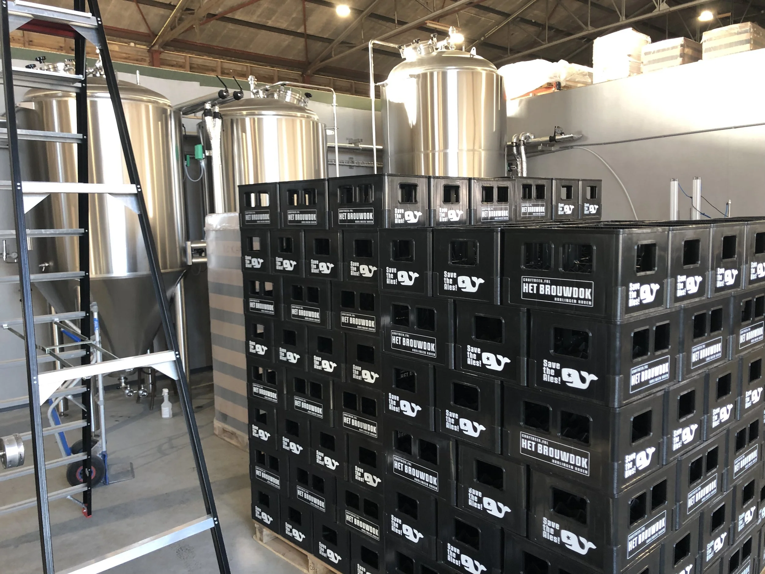 Industriële setting met grote metalen tanks, zwart houten kratten met tekst 'Het Brouwdok' en 'Save the Ales!', een metalen ladder en hoop dozen op een hoge plank. Geïndustrialiseerde brouwinstallatie.