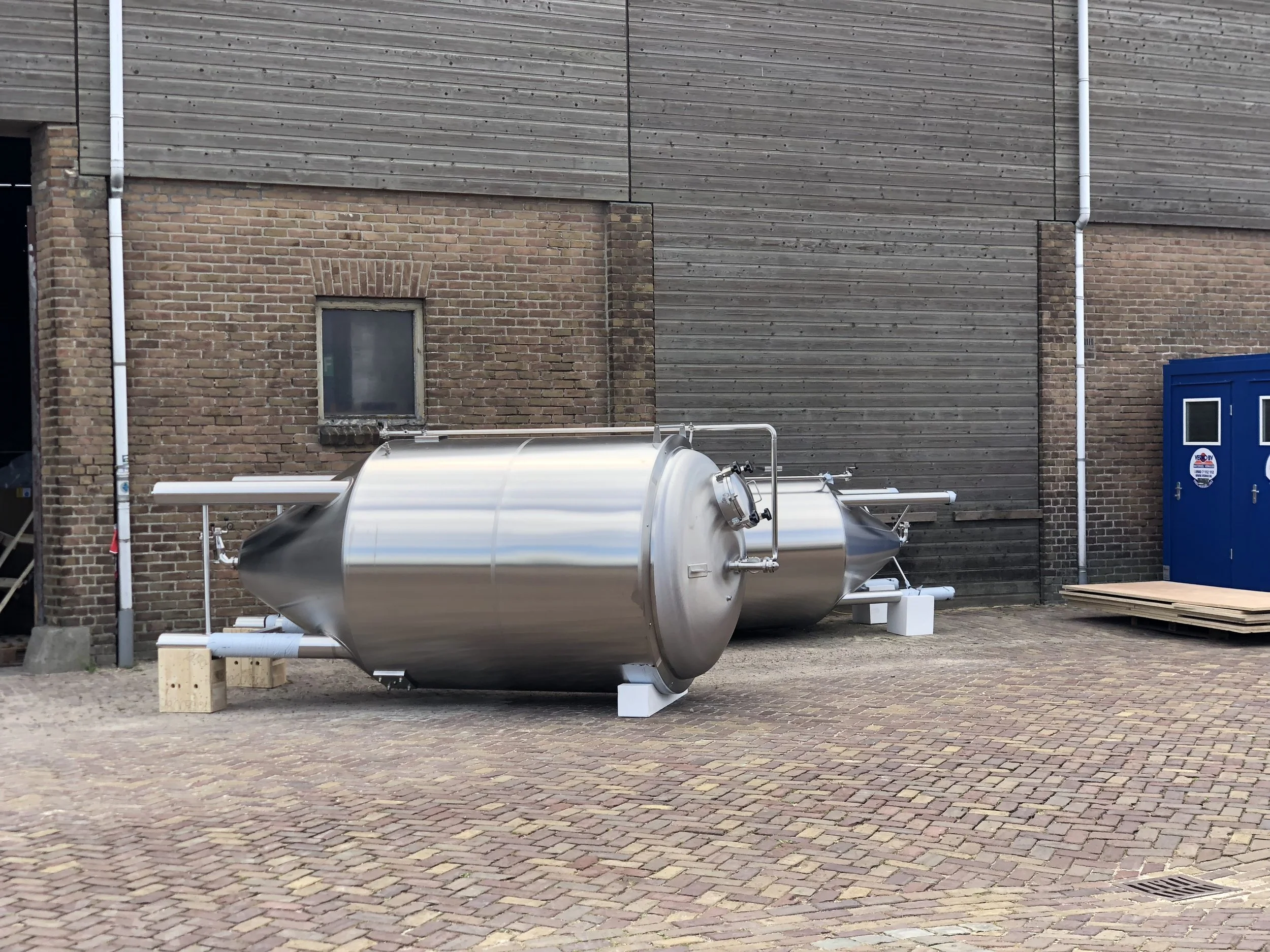 Een roestvrijstalen tank op een parkeerplaats met houten en bakstenen muren op de achtergrond. Kleine bouwwerkzaamheden en bouwmaterialen zijn zichtbaar.
