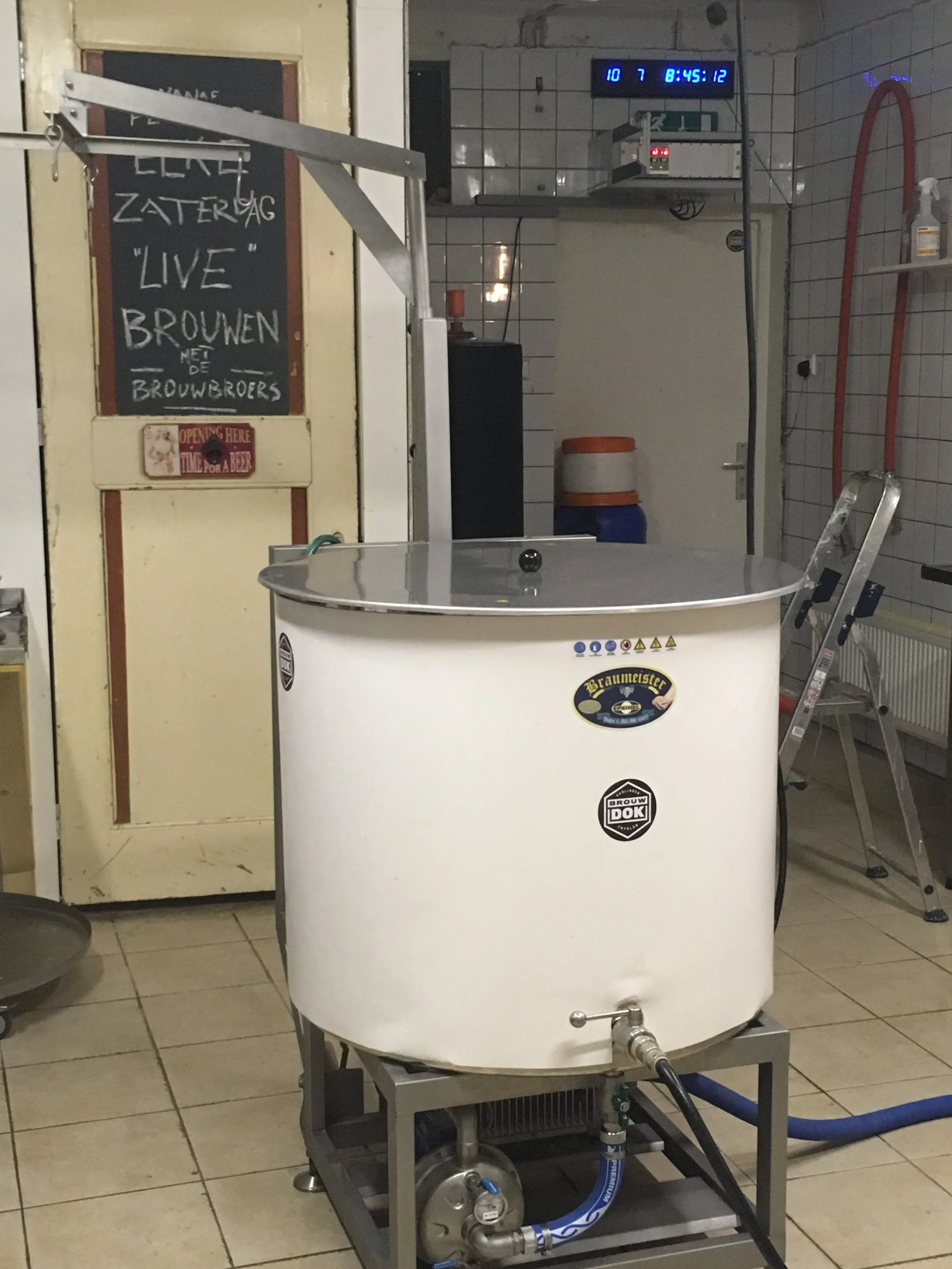 Een industrieel brewing apparaat met een cilindrisch vat en ingewikkelde pijpleidingen, in een omgeving met witte tegels en enkele herkenbare etiketten voor het brouwen van bier.