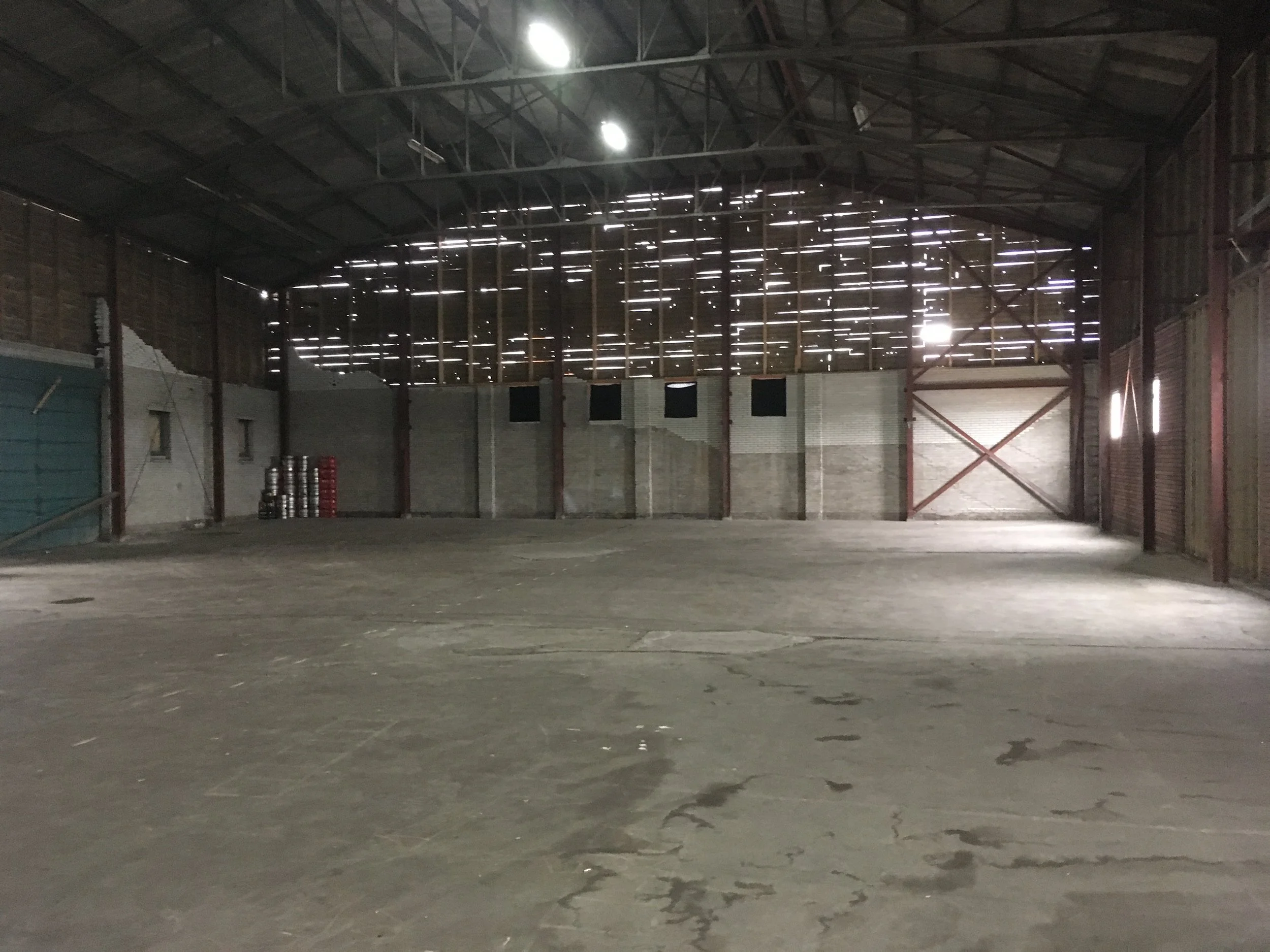 Binnenkant van een grote lege opslagloods met een houten plafond en metalen structuur, lichte gaatjes in het plafond laten licht door.
