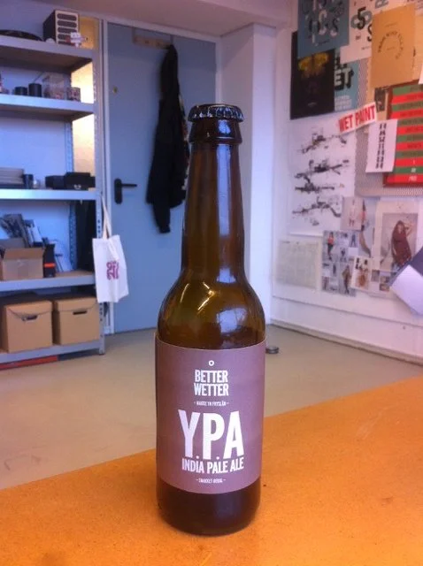 Een fles YPA India Pale Ale op een houten tafel in een interieur met boekenplanken en een muur met posters.