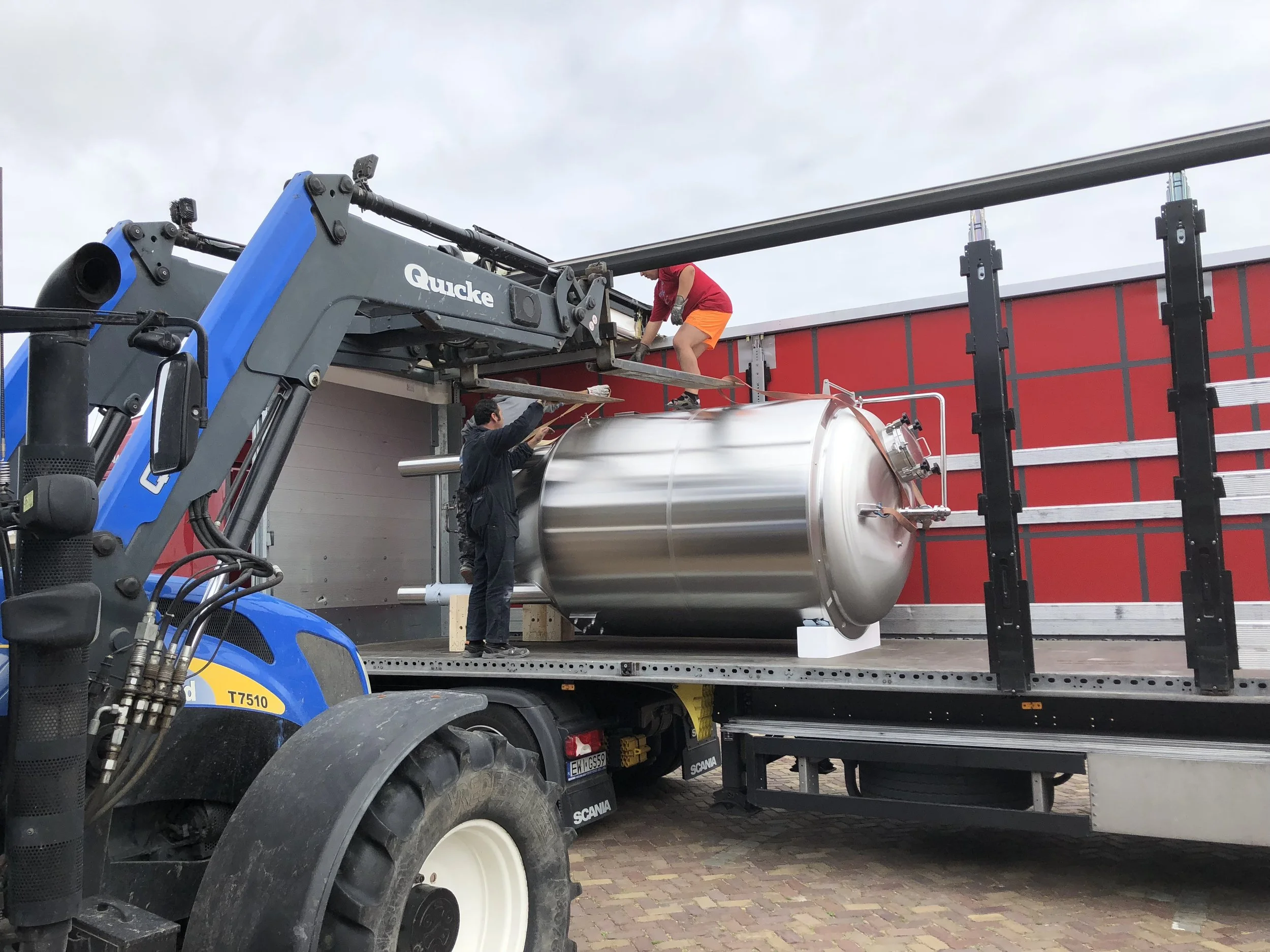 Mens in rode shirt en oranje korte broek staat op een ladder die op een vrachtwagen is gemonteerd, terwijl een andere man onderaan de ladder helpt bij het plaatsen van een grote, metalen tank op de vrachtwagen. Een heftruck staat op de voorgrond.