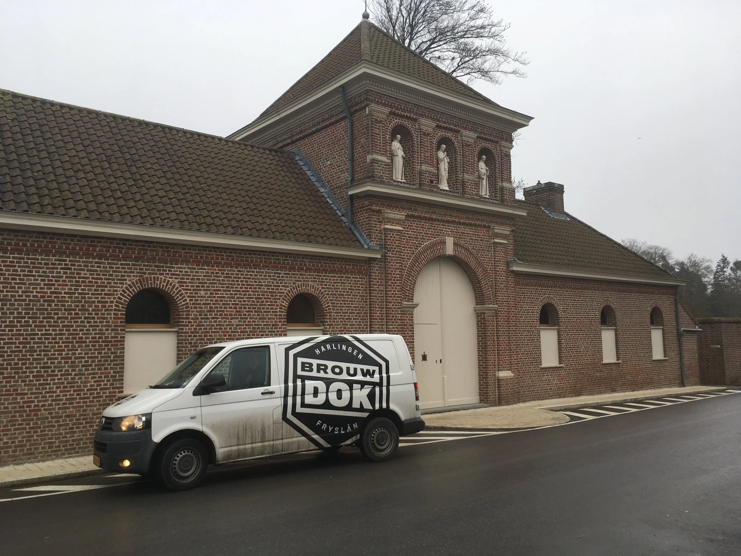 Een witte vrachtwagen met het logo 'Brouwer Fryslan' op de zijkant rijdt door een straat voor een oud bakstenen gebouw met drie standbeelden in nisjes op de bovenste verdieping.