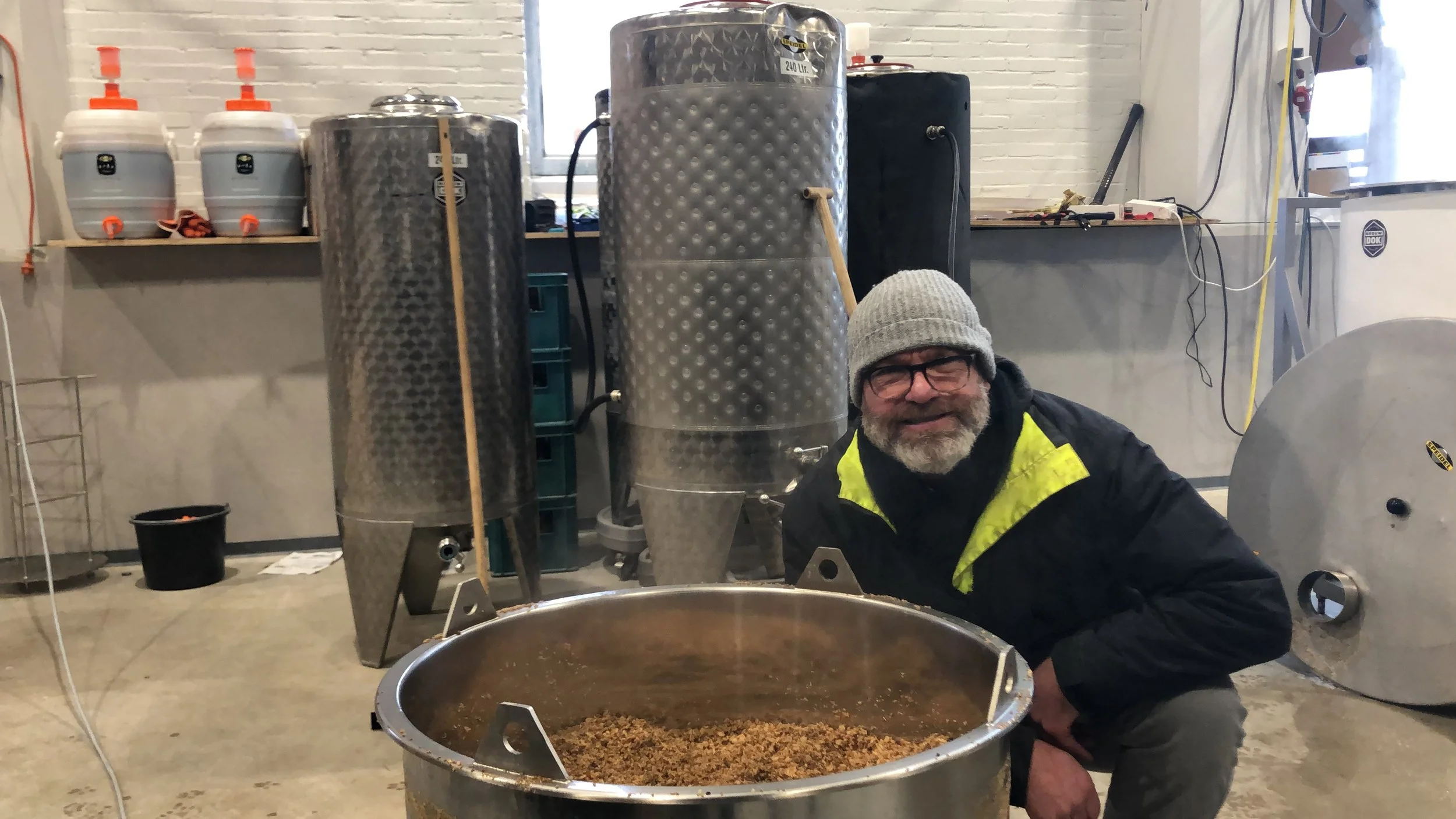 Man met bril, grijze muts en gele-zwart jasje die knielt naast grote metalfles met inhoud, mogelijk in een brouwerij of distilleerderij.