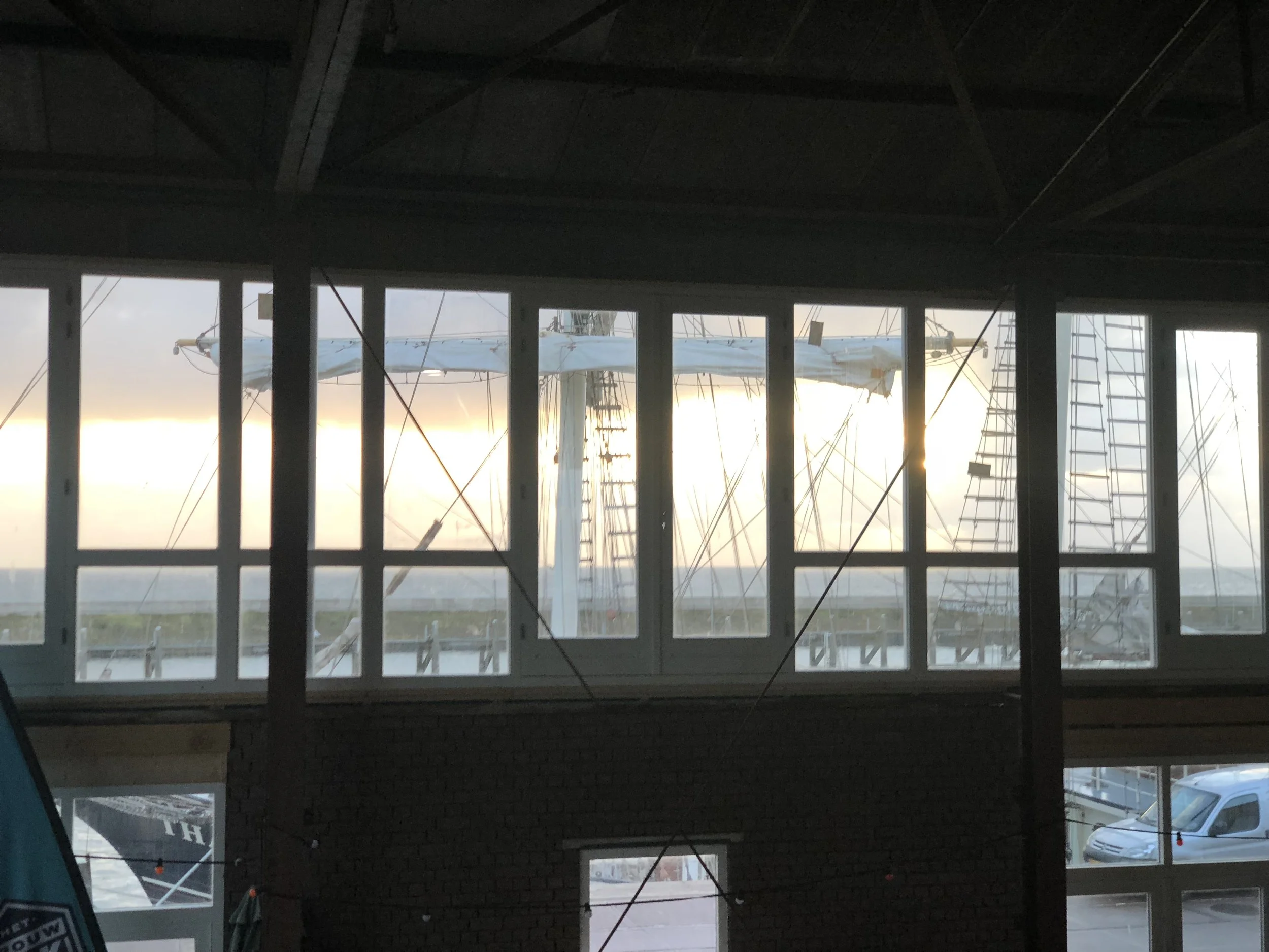 Binnenzicht van een gebouw onder constructie met grote ramen en een zeil tussen de spanten, met uitzicht op een bewolkte hemel en enkele voertuigen buiten.
