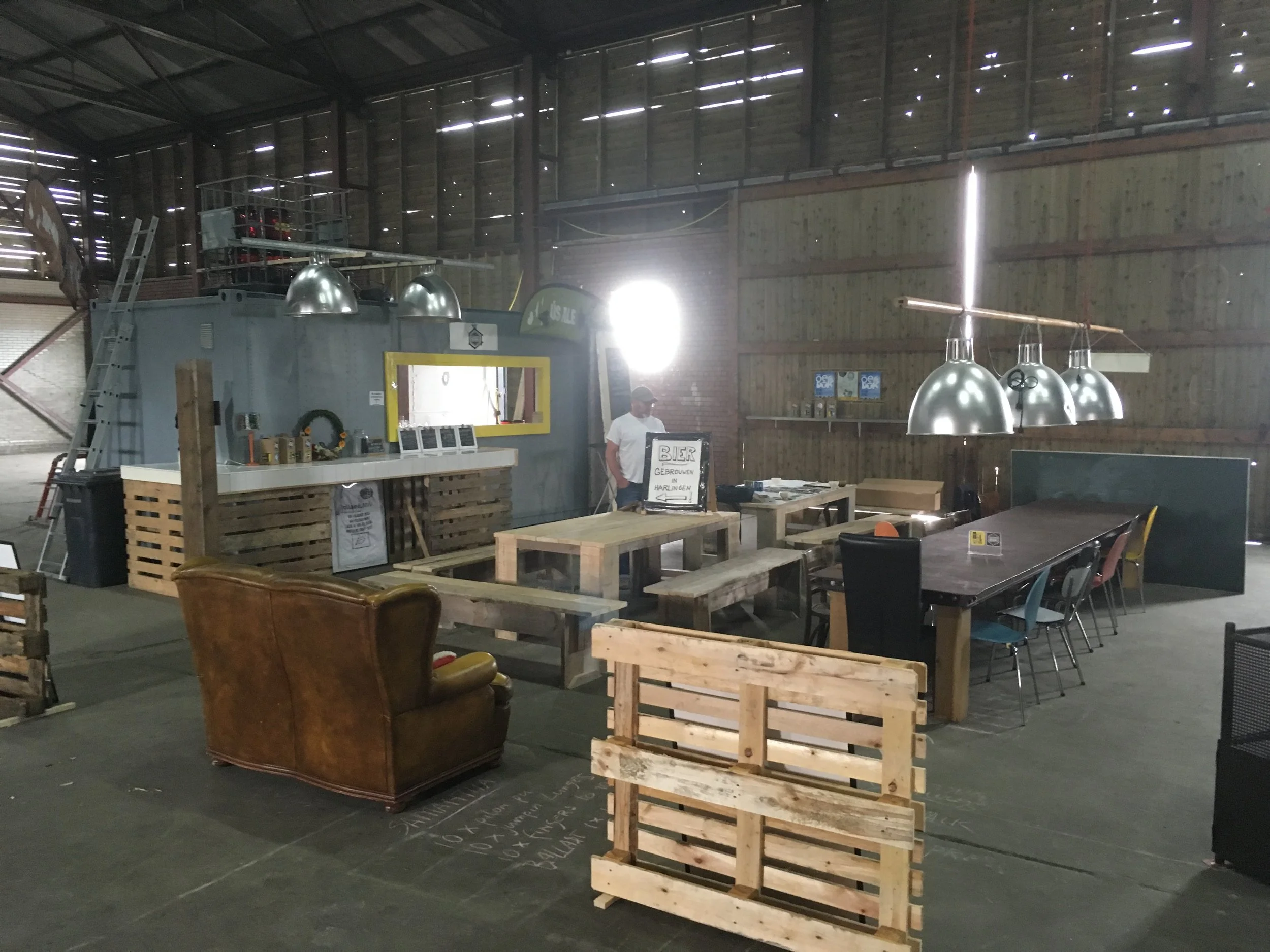 Binnenaanzicht van een ruimte met houten en metalen elementen, waaronder een bar, pallets, tafels, stoelen, hanglampen, en een persoon die op de achtergrond staat.