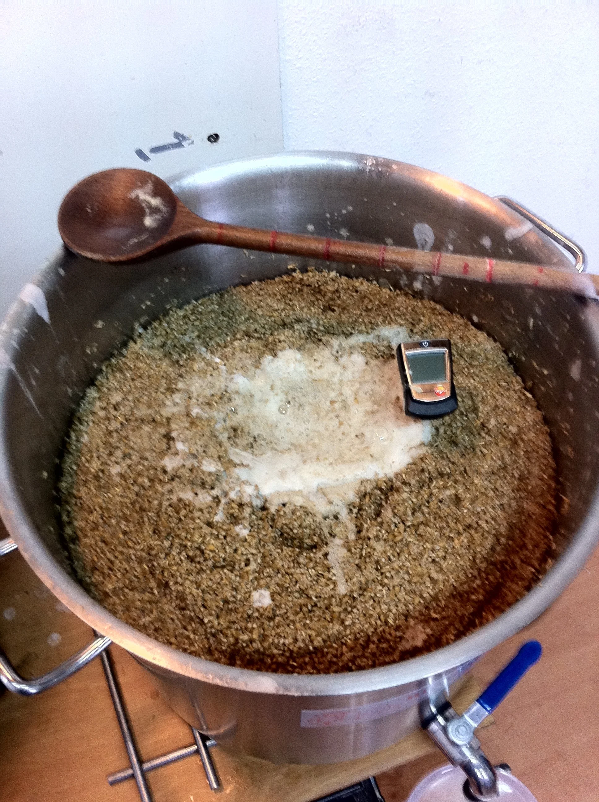 Een grote metalen machine waarin een mengsel van gemalen nootmuskaat en ander specerijen zit. Op de rand van de machine ligt een houten lepel en er liggen een pen en een klein apparaatje op de machine.