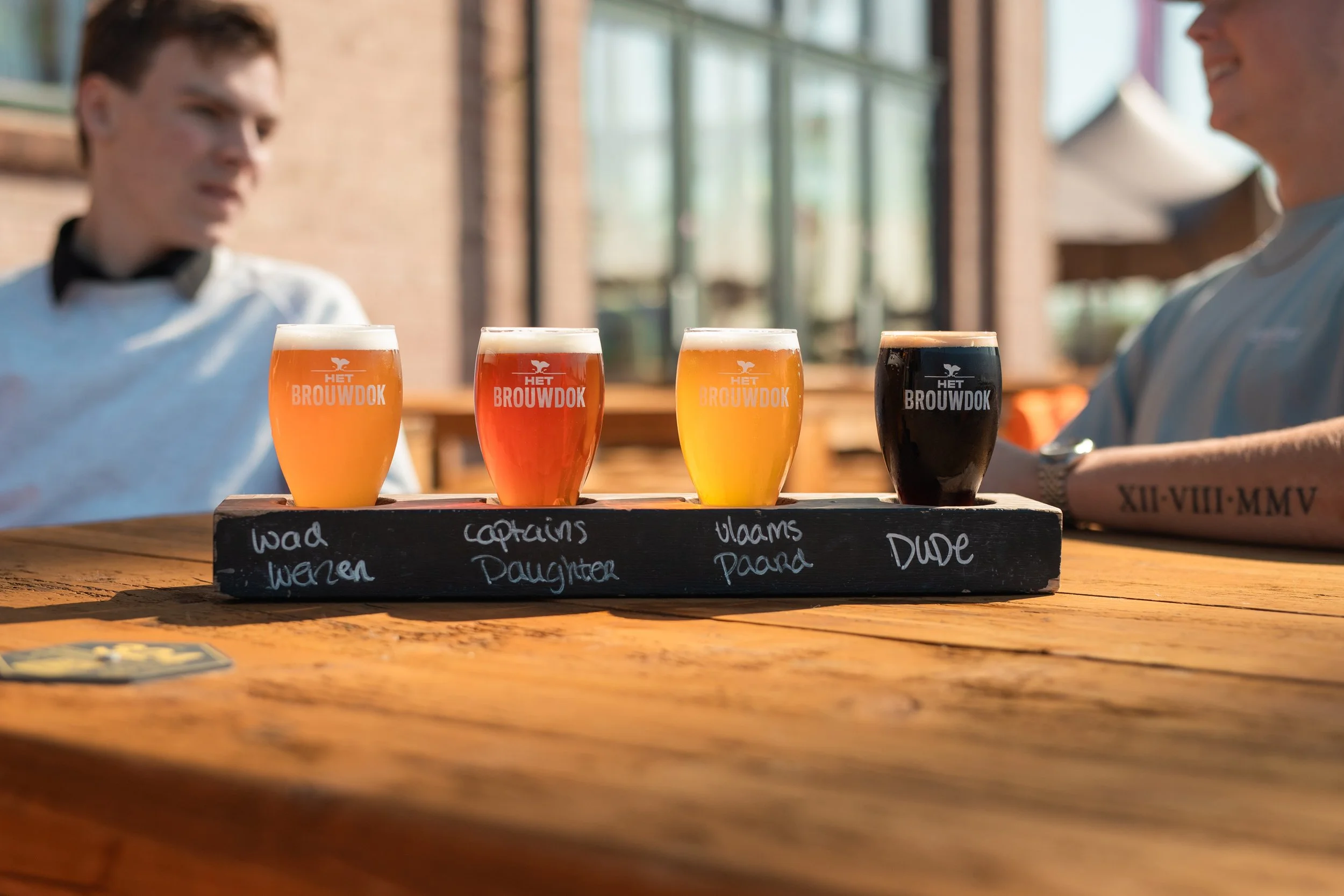 Vier verschillende soorten bieren in kleine glazen op een houten plank met schrift op een zwarte onderplaat, mensen zitten aan een tafel in een café of restaurant met grote ramen in de achtergrond.