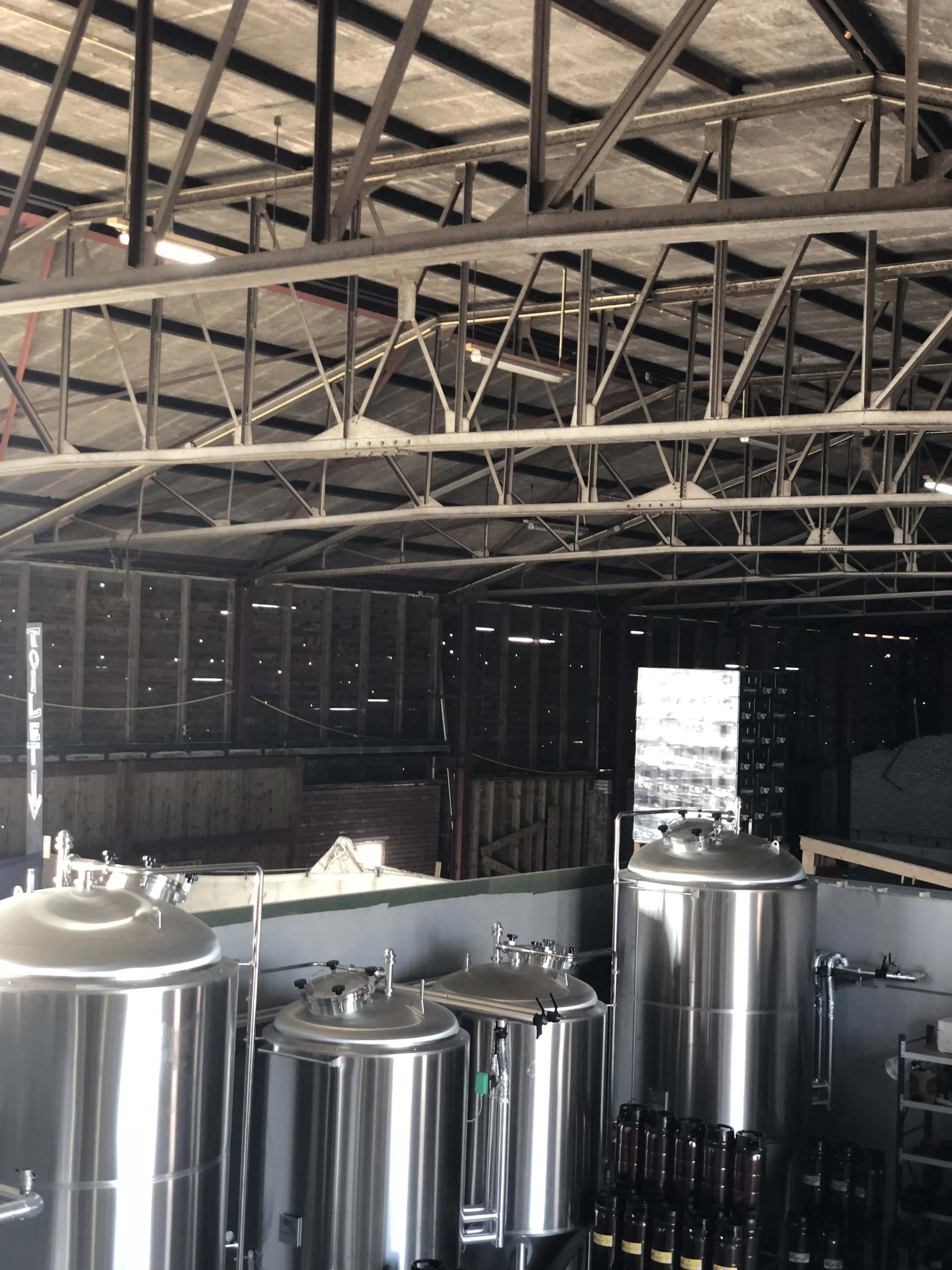 Een binnenaanzicht van een brouwerij met grote roestvrijstalen tanks, houten elementen en metalen trussen op het plafond.