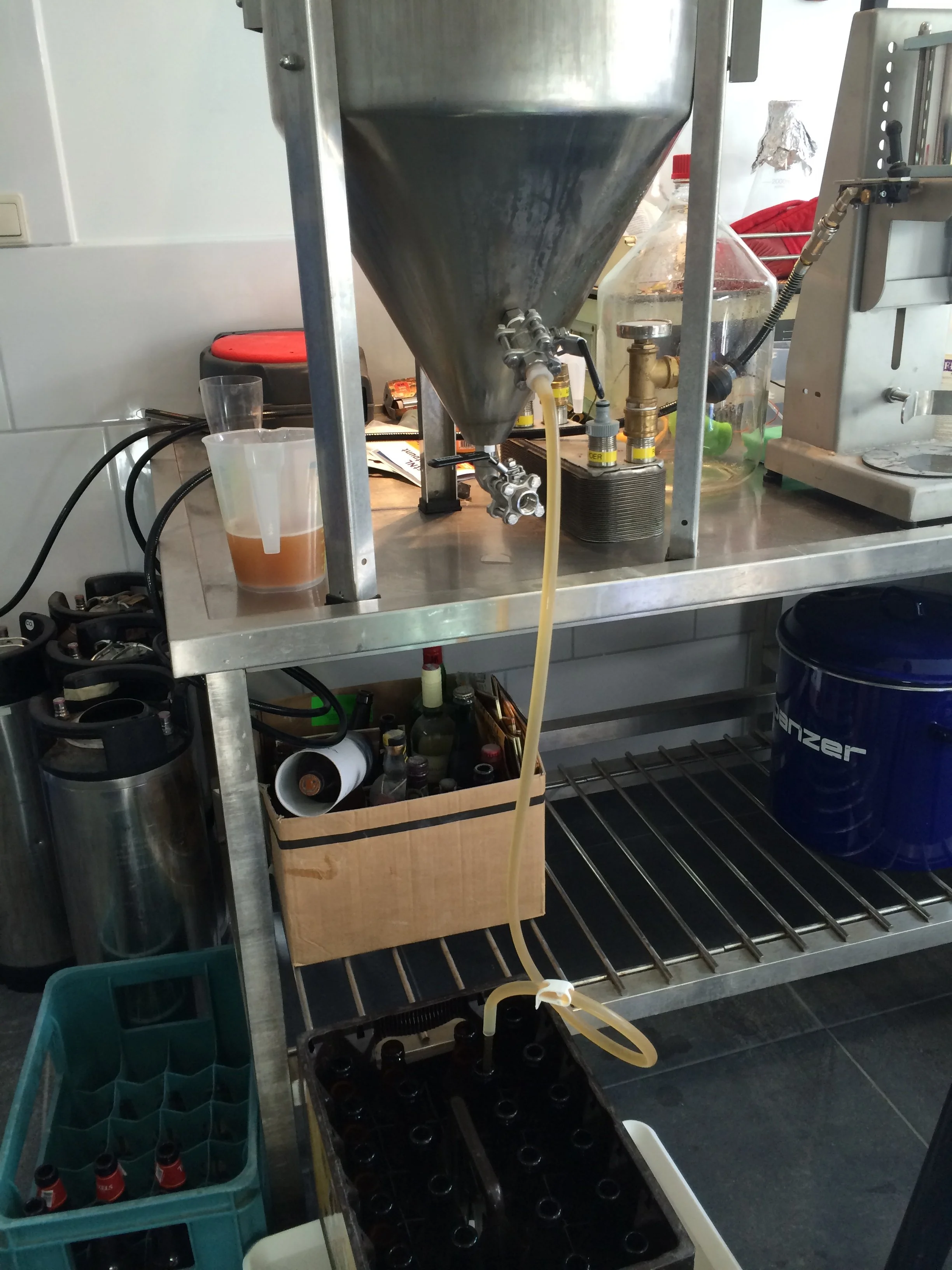 Een metaalwerkbank met chemische en laboratoriumapparatuur, een container met een amberkleurige vloeistof, en kisten met kleine flesjes en gekleurde doppen.