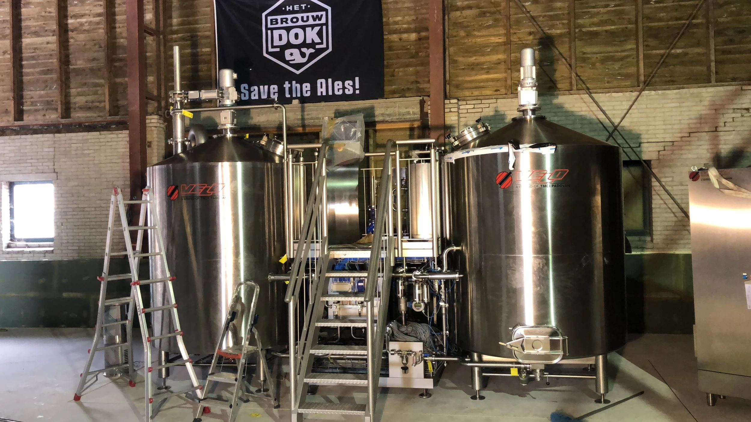 Twee grote, stalen tanks met leidingen en een ladder in een industriële ruimte, met een vlag met tekst 'Het Brouwdok Save the Ales!'