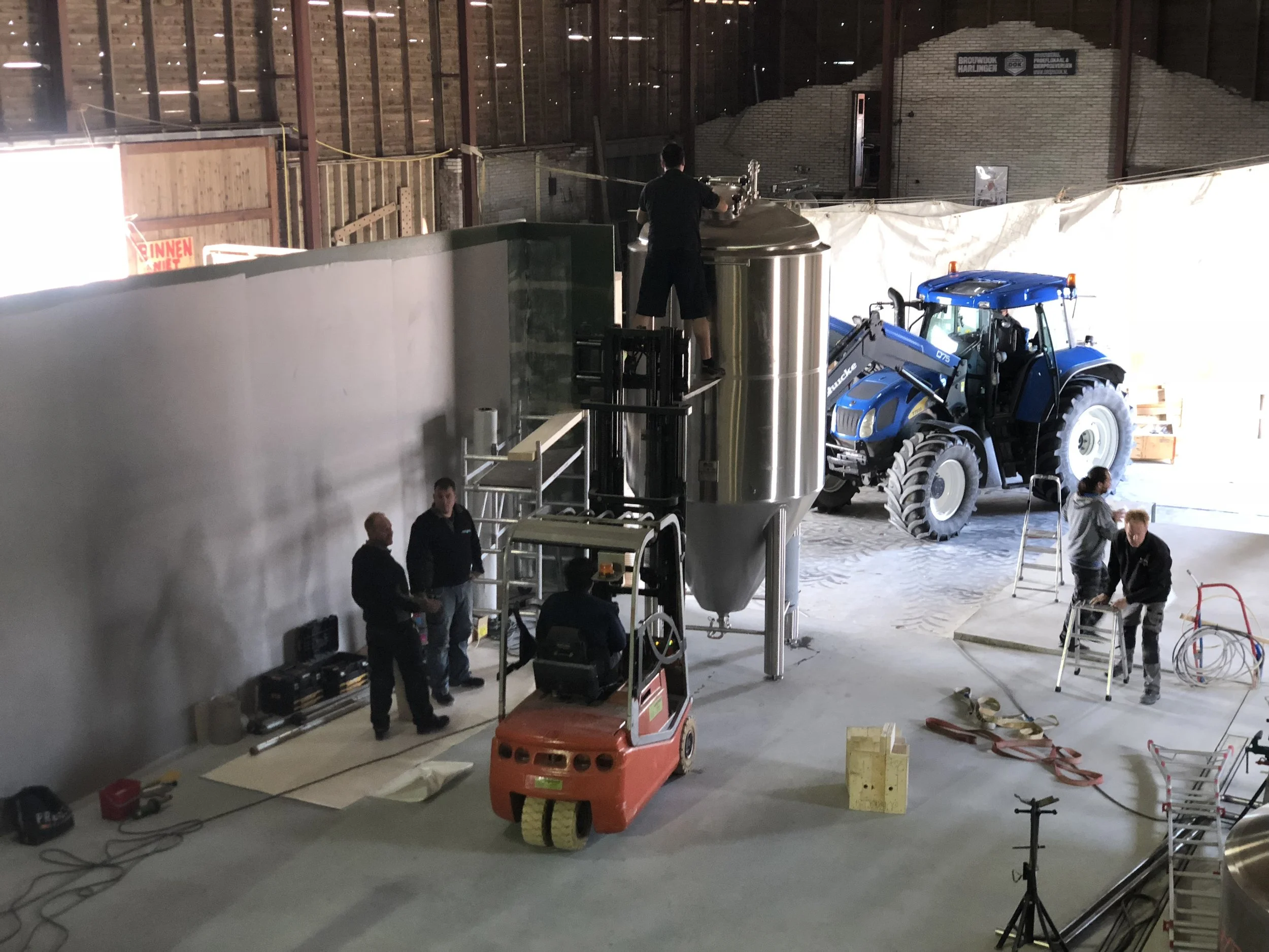 Een binnenruimte met mensen die bezig zijn met werkzaamheden. Er is een grote tractor, een grote metalen container, en verschillende bouwmaterialen en gereedschappen.