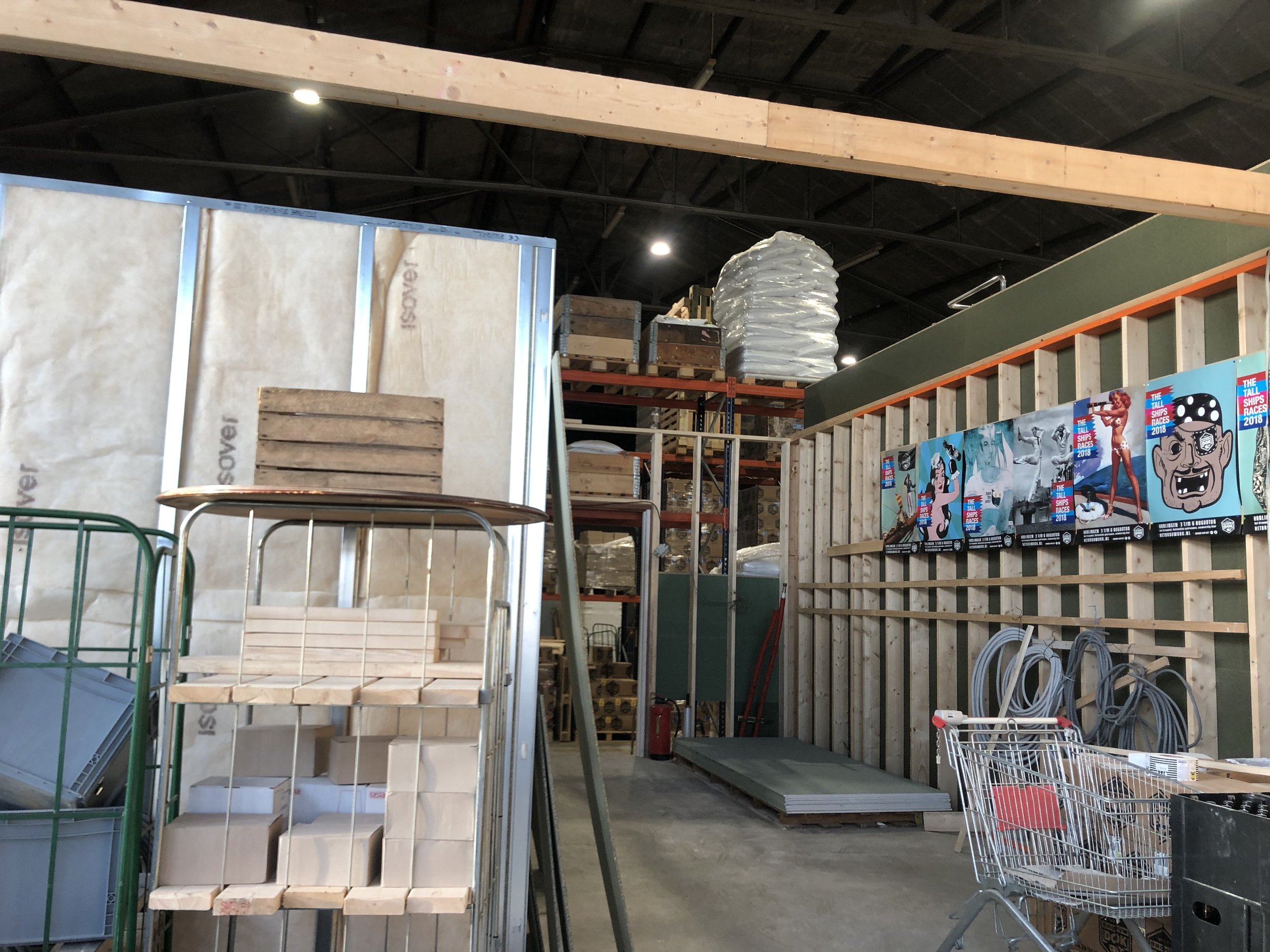 Kassawagen in een bouwmarkt met winkelwagentje, houten planken en bouwmaterialen op een metalen rek, binnen een magazijn met hoge plafonds en verlichtingslichten.