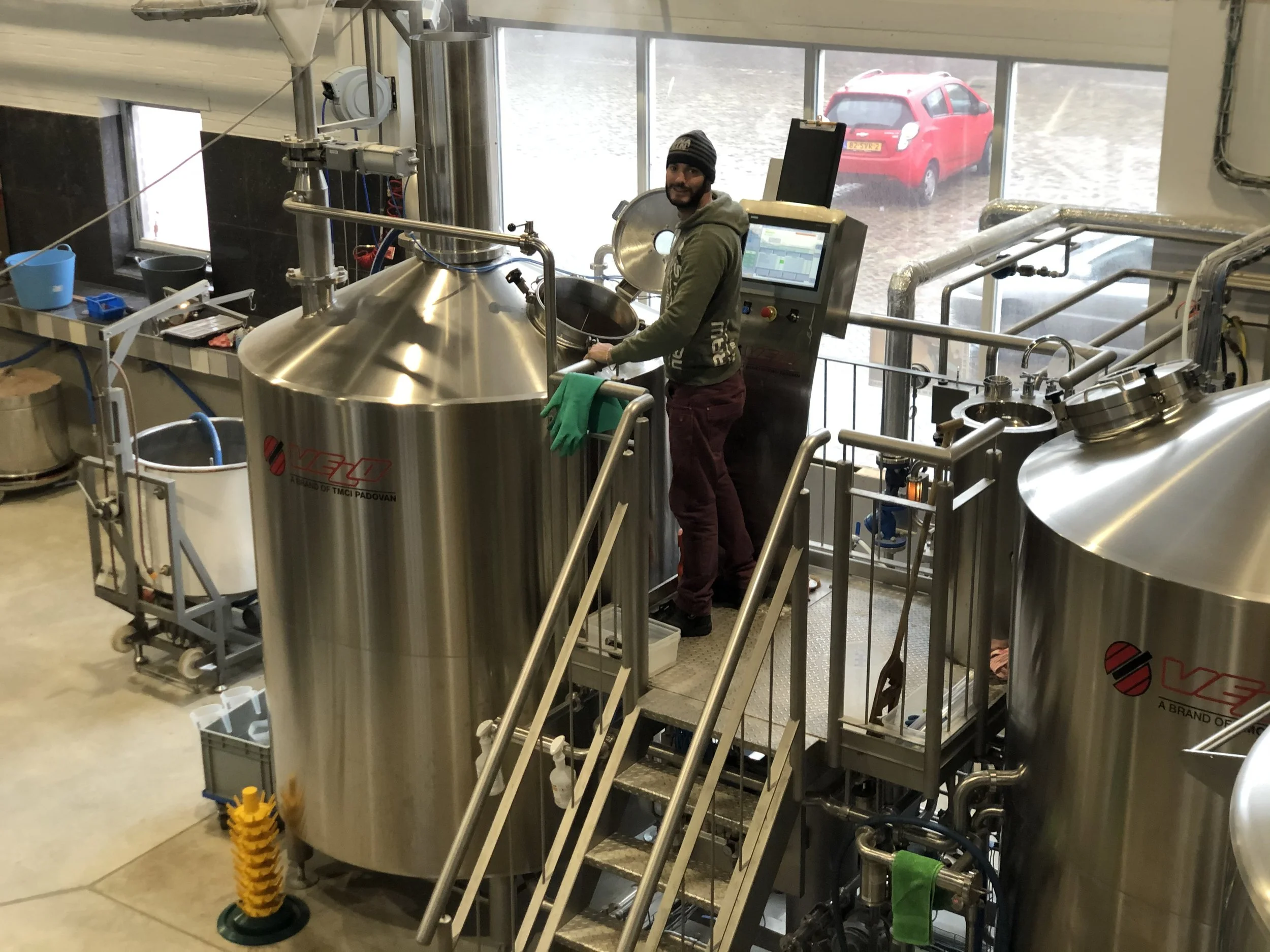 Een persoon die in een brouwerijbrouwinstallatie werkt, omringd door roestvrijstalen tanks, met een computerscherm en een grote raam met uitzicht op een rood auto buiten.